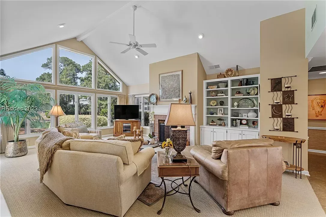 A bright living room with a vaulted ceiling and floor-to-ceiling windows features a brick fireplace flanked by white built-in shelving, leather and upholstered seating, and a ceiling fan overhead.