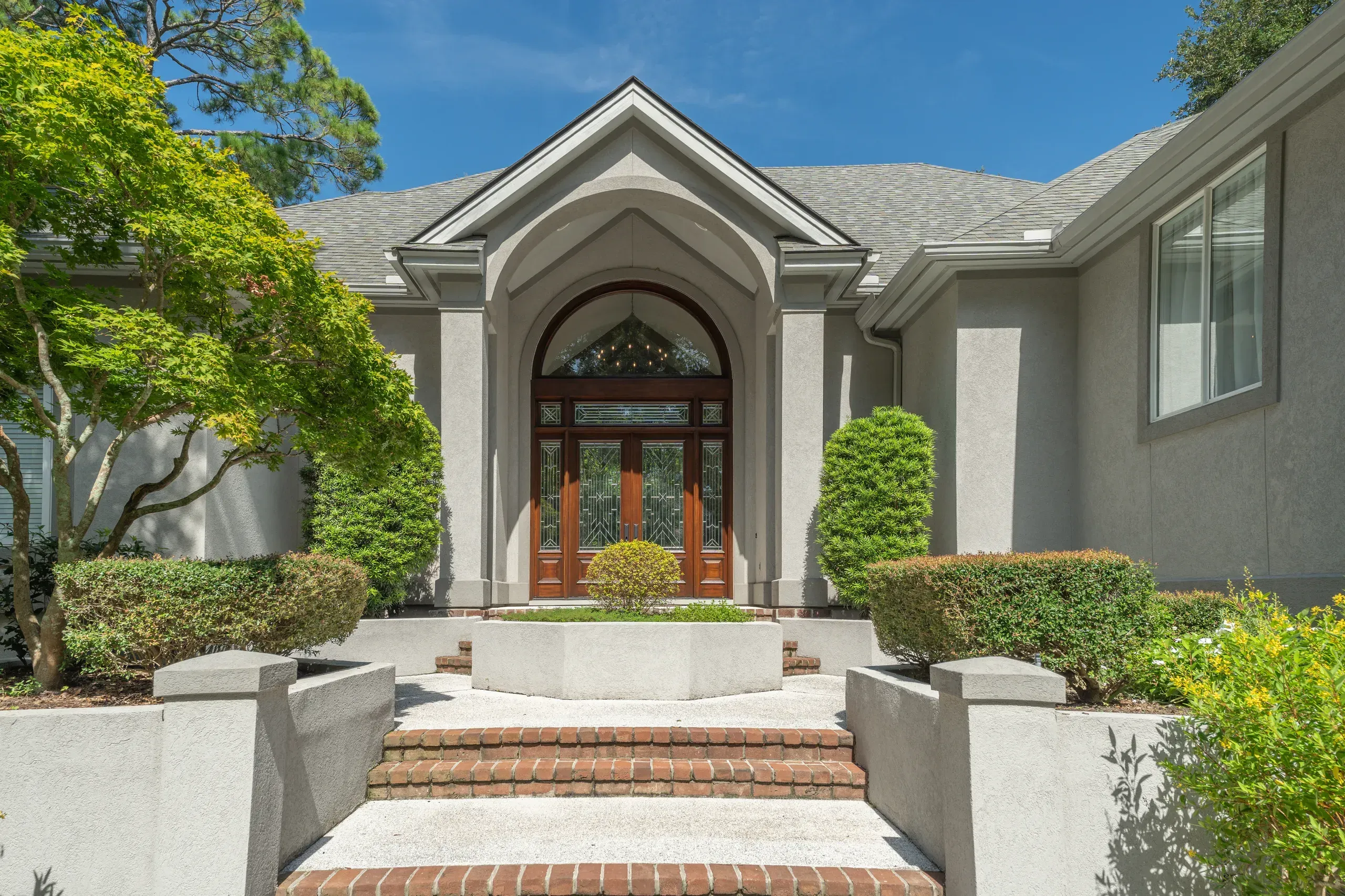 A stucco home entrance with a tall arched wooden double door topped by a fanlight window sits beneath a peaked gable roof, approached by brick steps and flanked by manicured shrubs and trees.