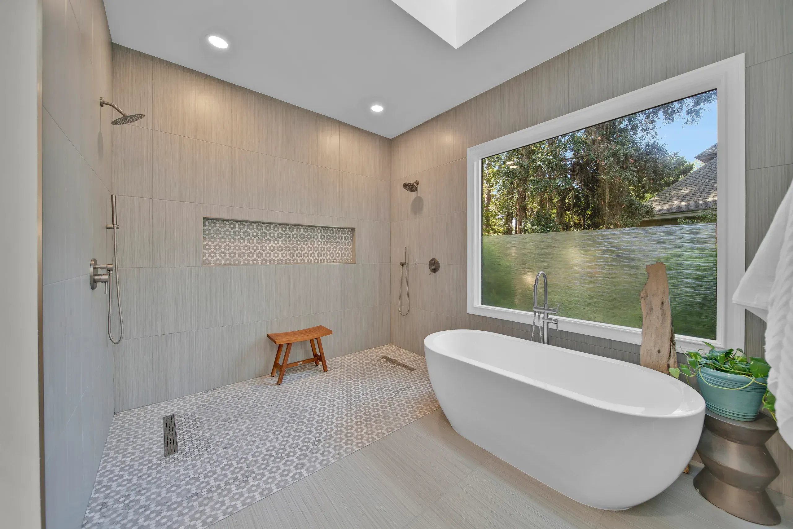 A spa-like bathroom remodel features an oval freestanding tub beside a large window overlooking trees, with an open curbless tile shower and teak bench in the background.