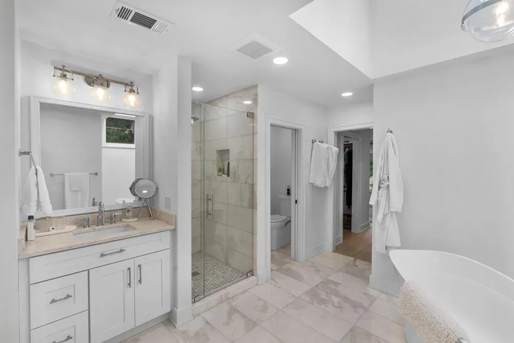 A spacious primary bathroom features a white vanity with marble-look countertop, a glass-enclosed tiled shower, a freestanding soaking tub, and a separate water closet, all lit by a skylight and warm sconce lighting.