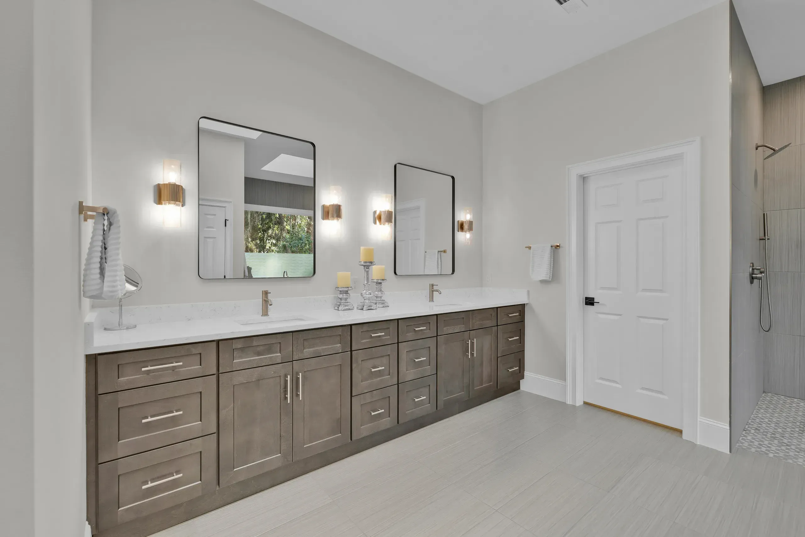 A primary bathroom features a long dark wood double vanity with white quartz countertops, two rectangular mirrors flanked by brass sconces, and a tiled walk-in shower visible at the far right.
