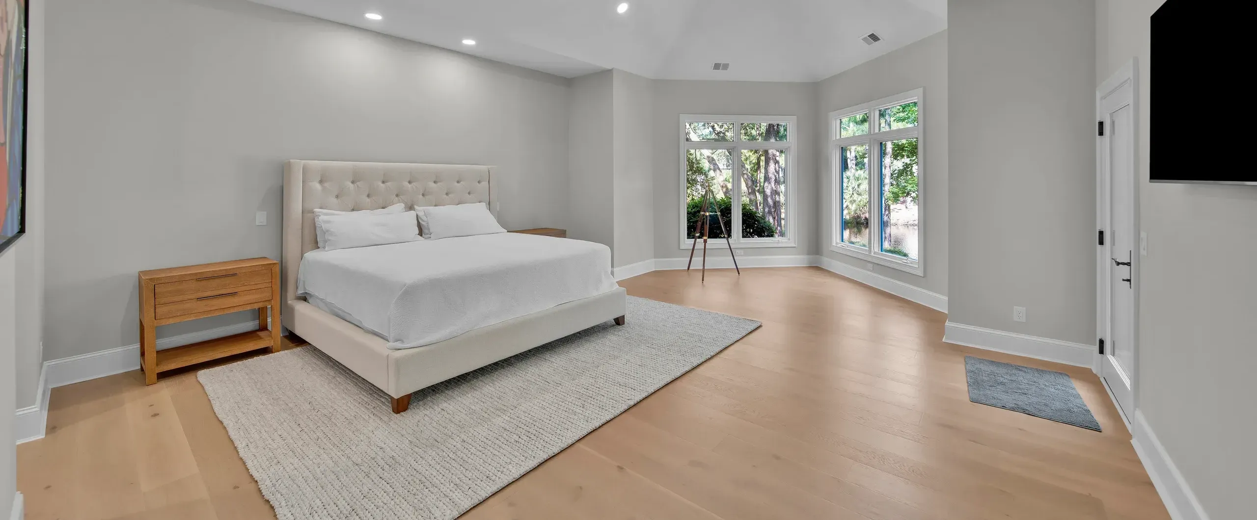 A spacious primary bedroom with a vaulted ceiling, a wood-bladed ceiling fan, an upholstered king bed on a light rug, and wide windows overlooking trees.