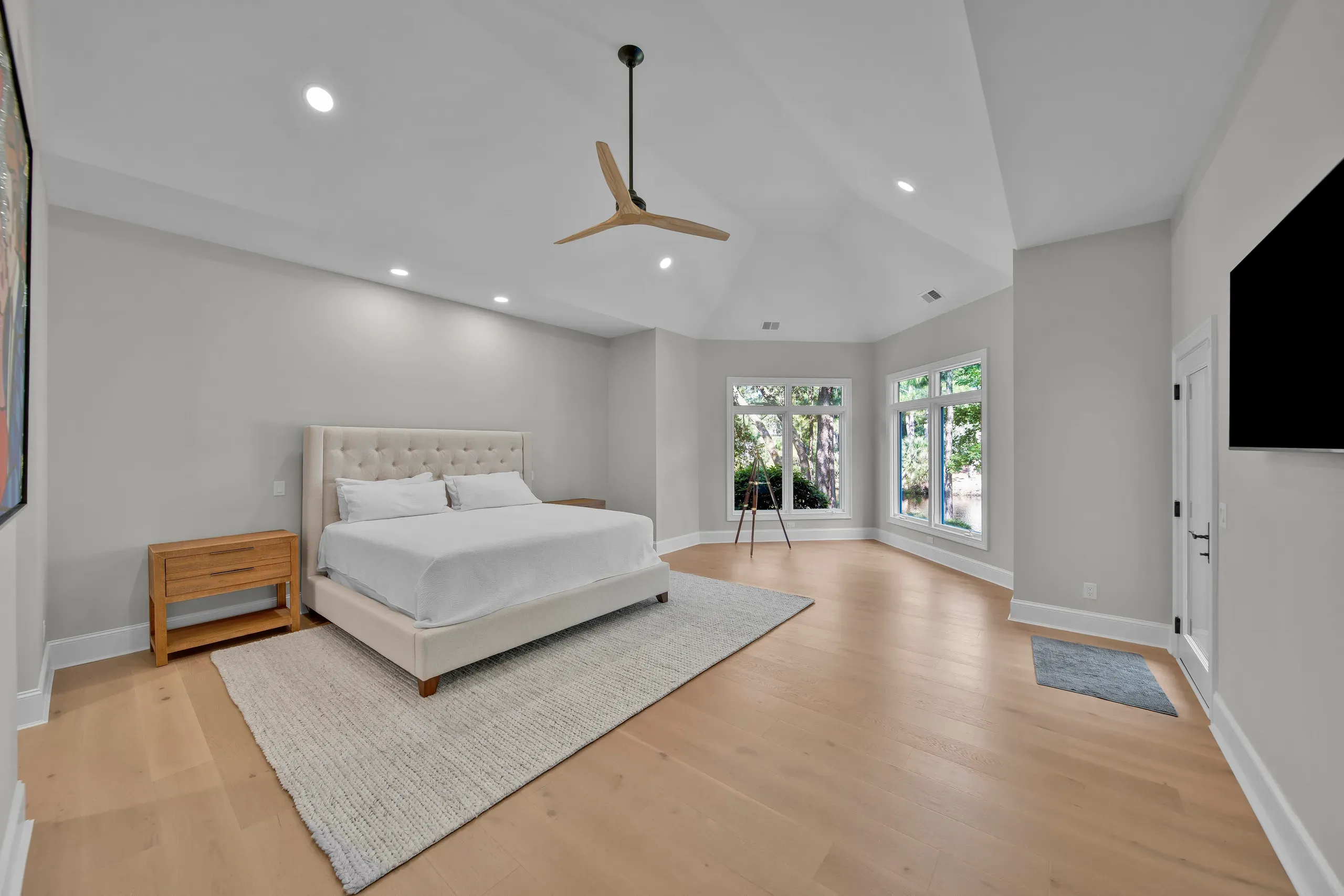 A spacious primary bedroom with a vaulted ceiling, a wood-bladed ceiling fan, an upholstered king bed on a light rug, and wide windows overlooking trees.
