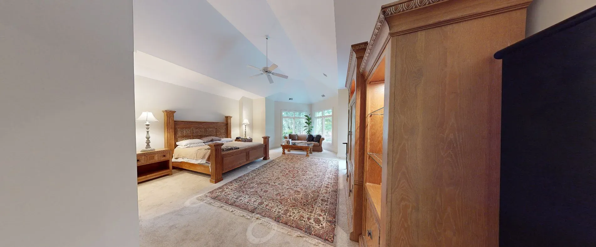A spacious primary bedroom with a vaulted ceiling features a carved wood bed and armoire, a patterned area rug over carpet, and a sitting area with sofa and coffee table near large windows.