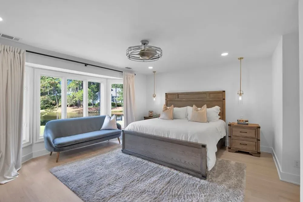 A primary bedroom with a wood-framed bed flanked by hanging pendant lights and a plush gray area rug, with a bay window offering a view of a lake and trees beyond a blue upholstered bench.