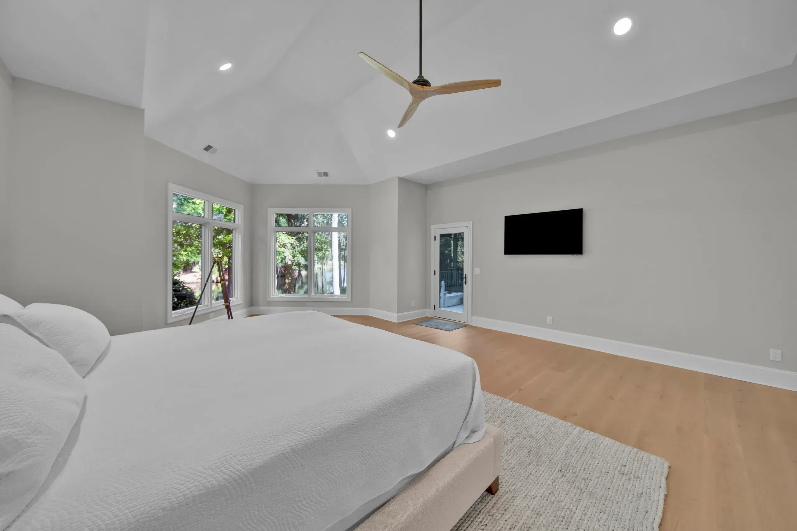A spacious primary bedroom with a vaulted ceiling, light wood flooring, a wall-mounted television, and large windows overlooking greenery beside a white upholstered bed.