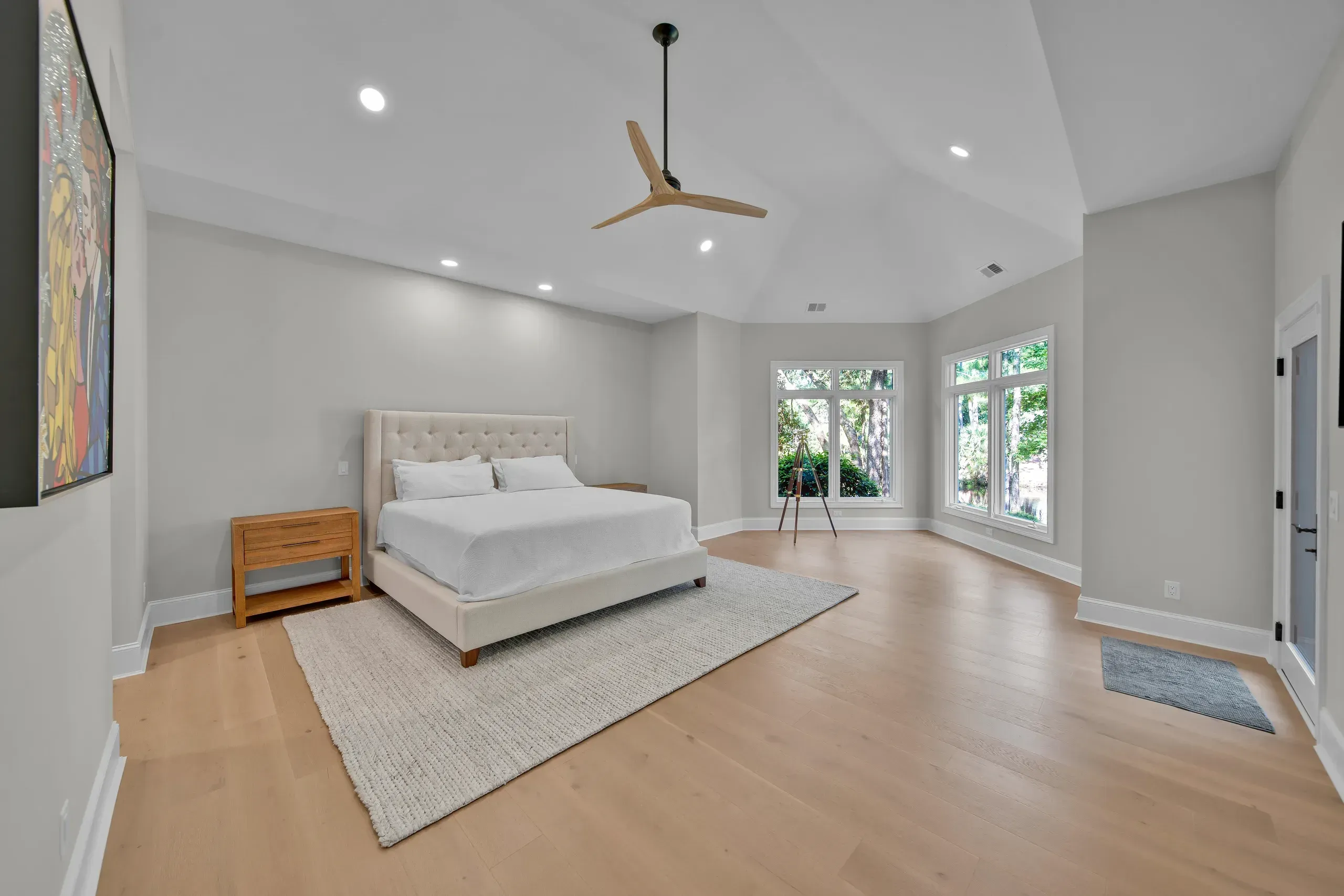 A spacious primary bedroom with a vaulted ceiling, upholstered bed, wood-plank flooring, and large windows overlooking trees.