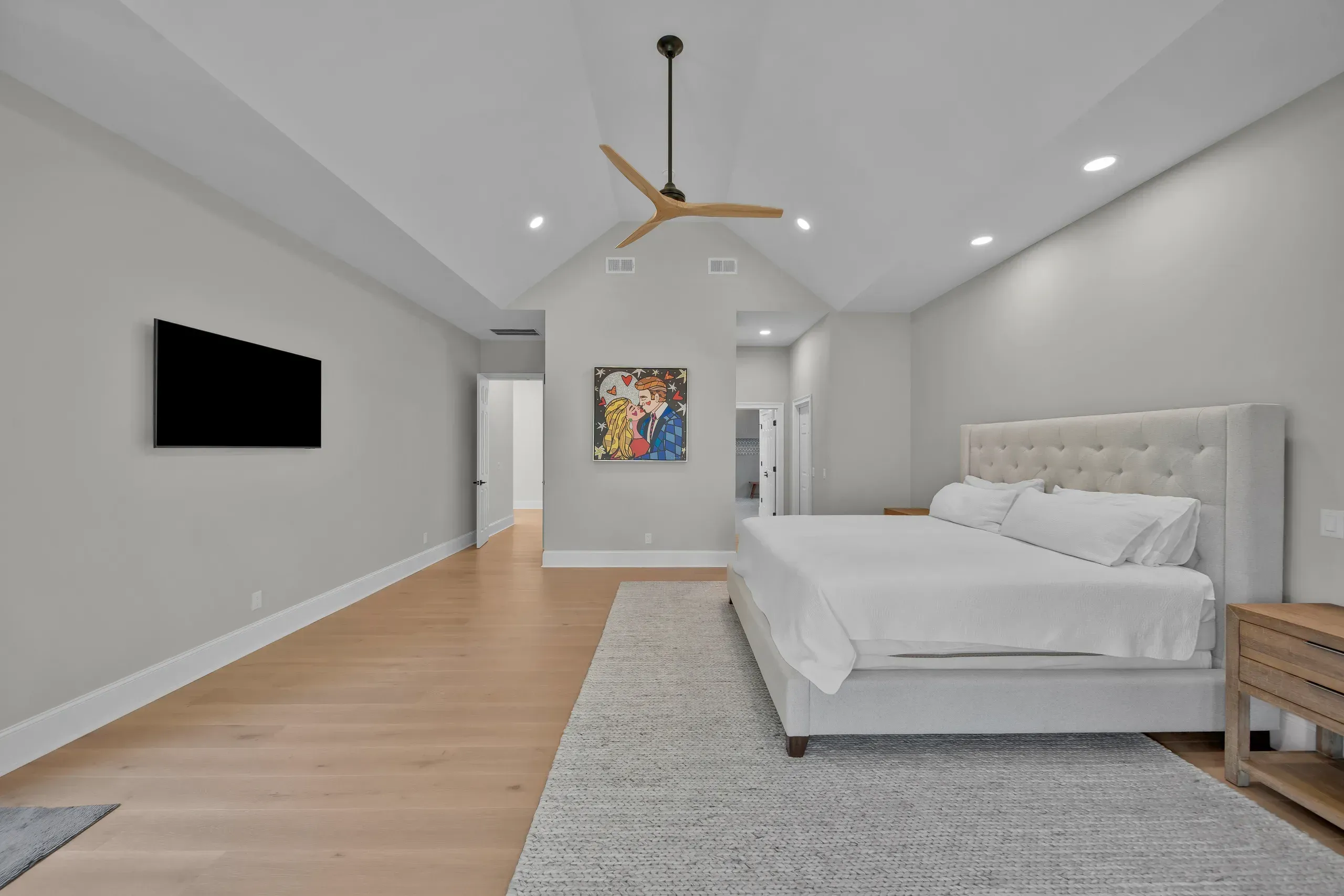 A spacious primary bedroom features a vaulted ceiling with a wood-bladed fan, a tufted upholstered bed, wide-plank wood flooring, and a hallway leading to an ensuite bathroom.