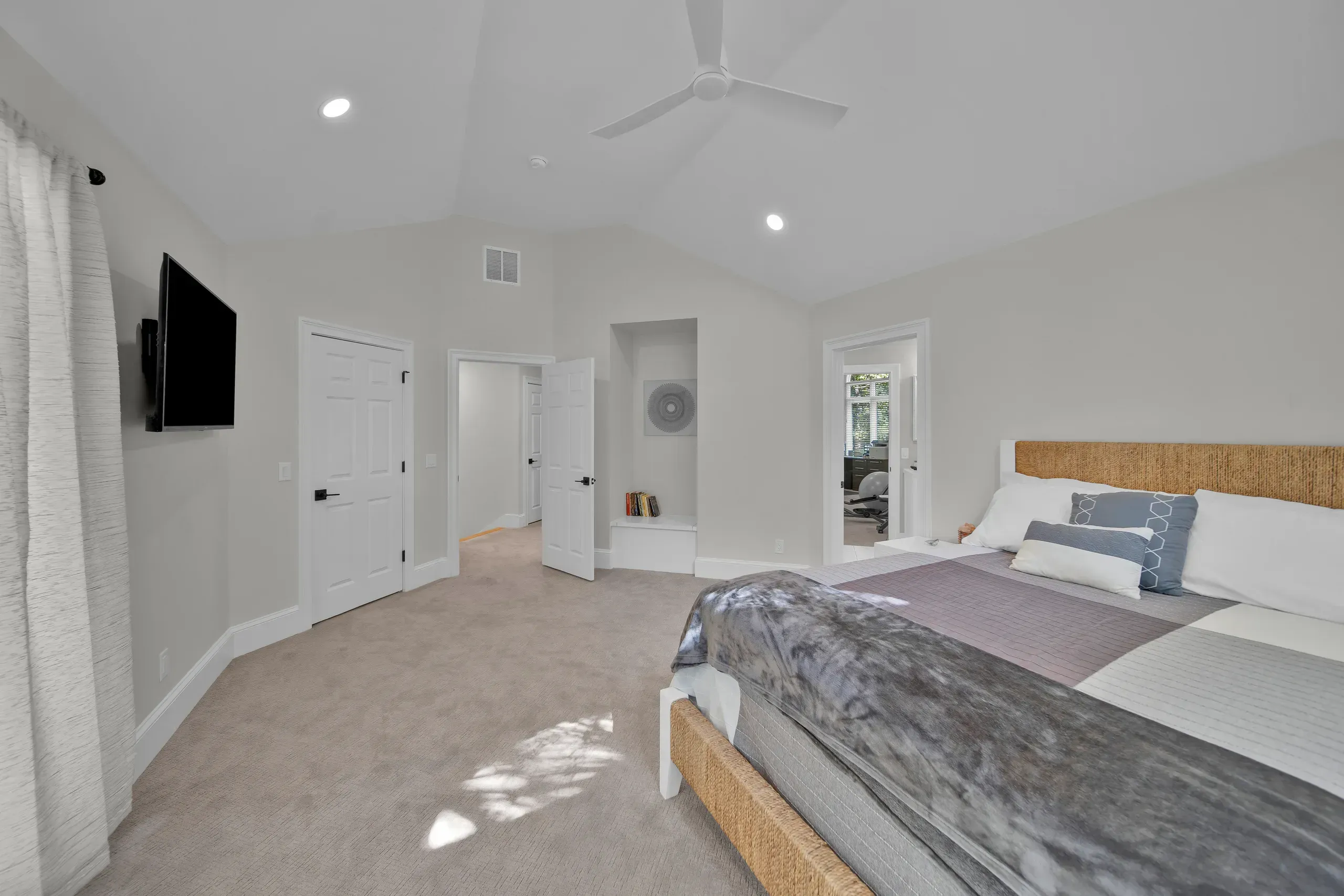 A spacious primary bedroom features a vaulted ceiling with a ceiling fan, plush gray carpet, a woven-headboard bed, a wall-mounted television, and a doorway leading to an adjoining home gym.
