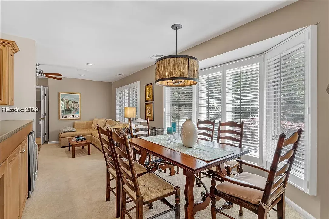 An open dining and living area features a wooden table with ladder-back chairs beneath a drum pendant light, with a bay window and plantation shutters filling the room with natural light.