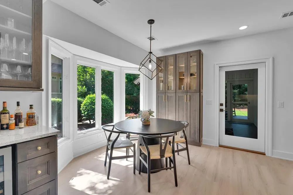 A round dining table with wishbone chairs sits in a bay-windowed breakfast nook beside a built-in wood cabinet with glass display doors and a geometric pendant light overhead, with a glass door leading outside to the right.