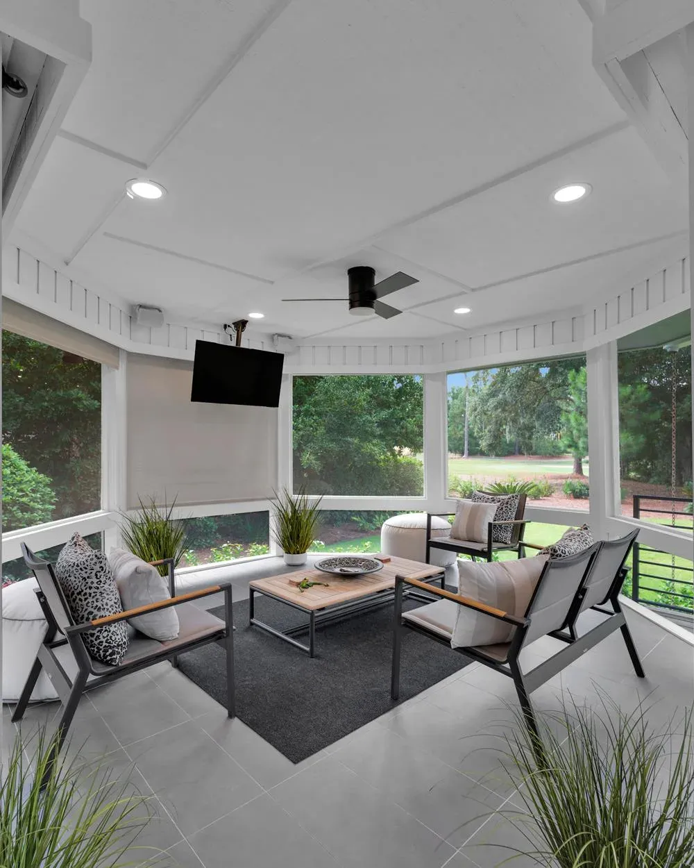 A screened porch addition features a wall-mounted television, ceiling fan, and modern outdoor seating arranged around a wooden coffee table with a golf course visible through the screens.
