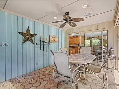 A screened porch with light blue wood plank walls features a ceiling fan, a metal star decoration, a tile floor, and a glass table with patio chairs beside a sliding door leading to the kitchen.