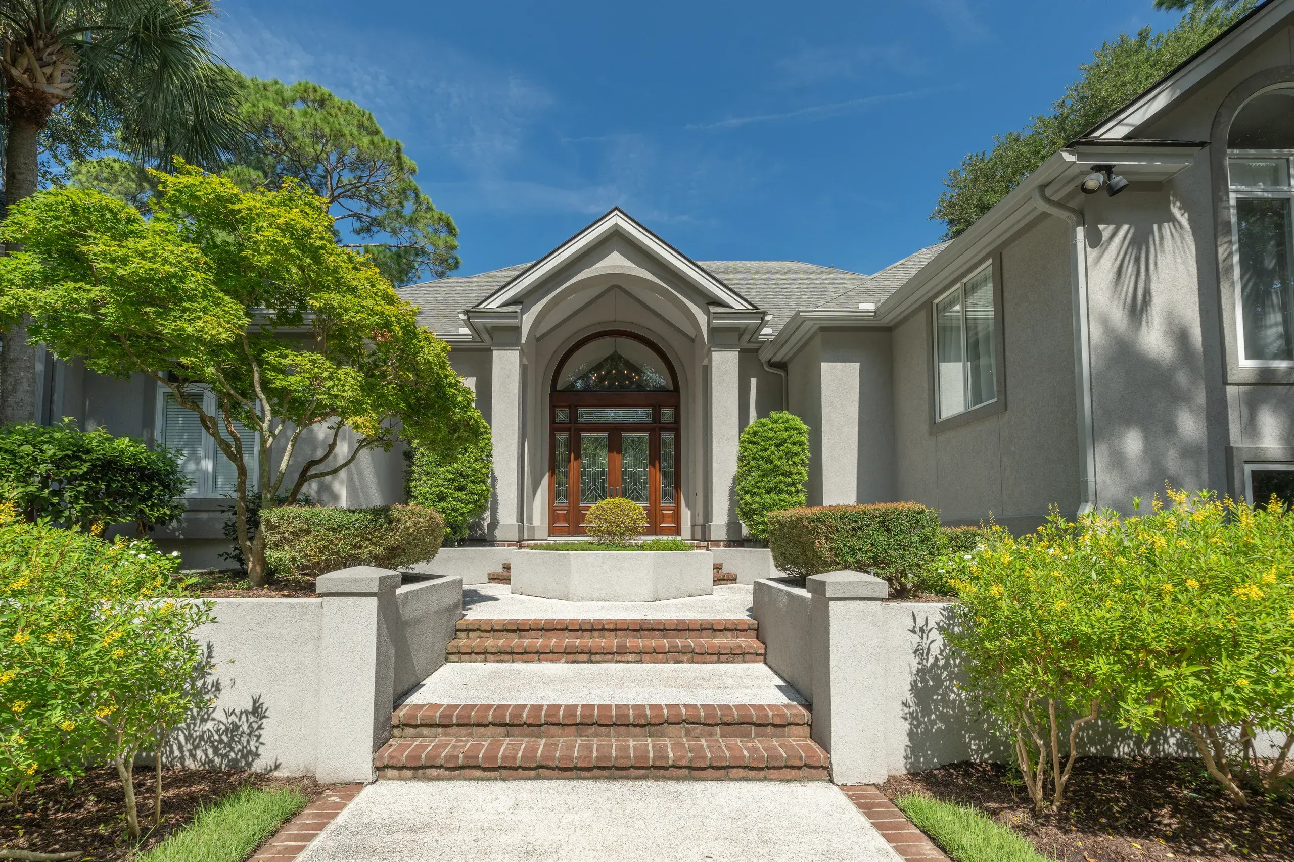 A stucco home entrance with an arched wooden double door beneath a tall gabled portico, approached by a brick-and-concrete staircase flanked by manicured shrubs and trees.