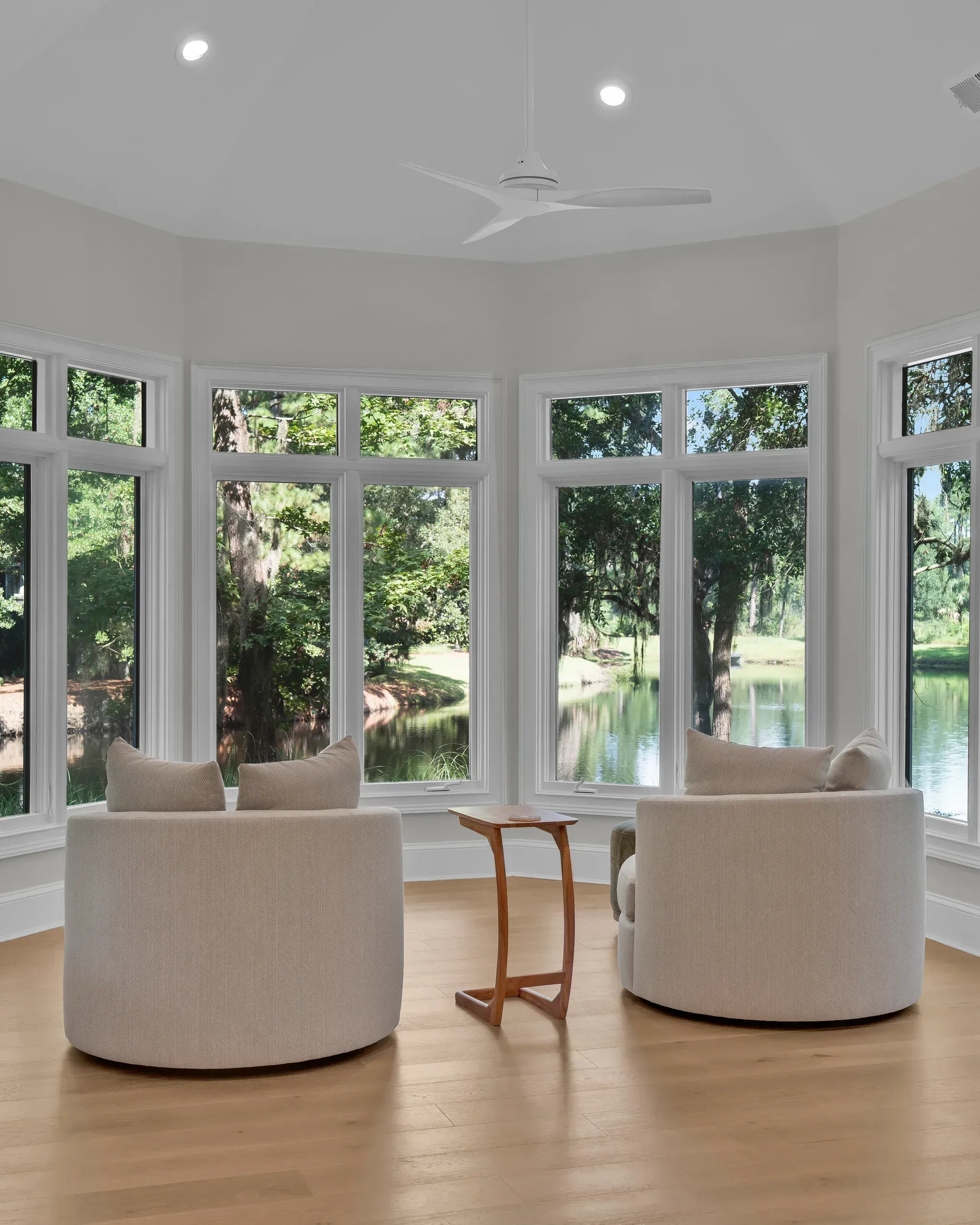 A bright sunroom with wraparound windows overlooking a lagoon features two round upholstered chairs, a small wooden side table, a ceiling fan, and warm hardwood flooring.