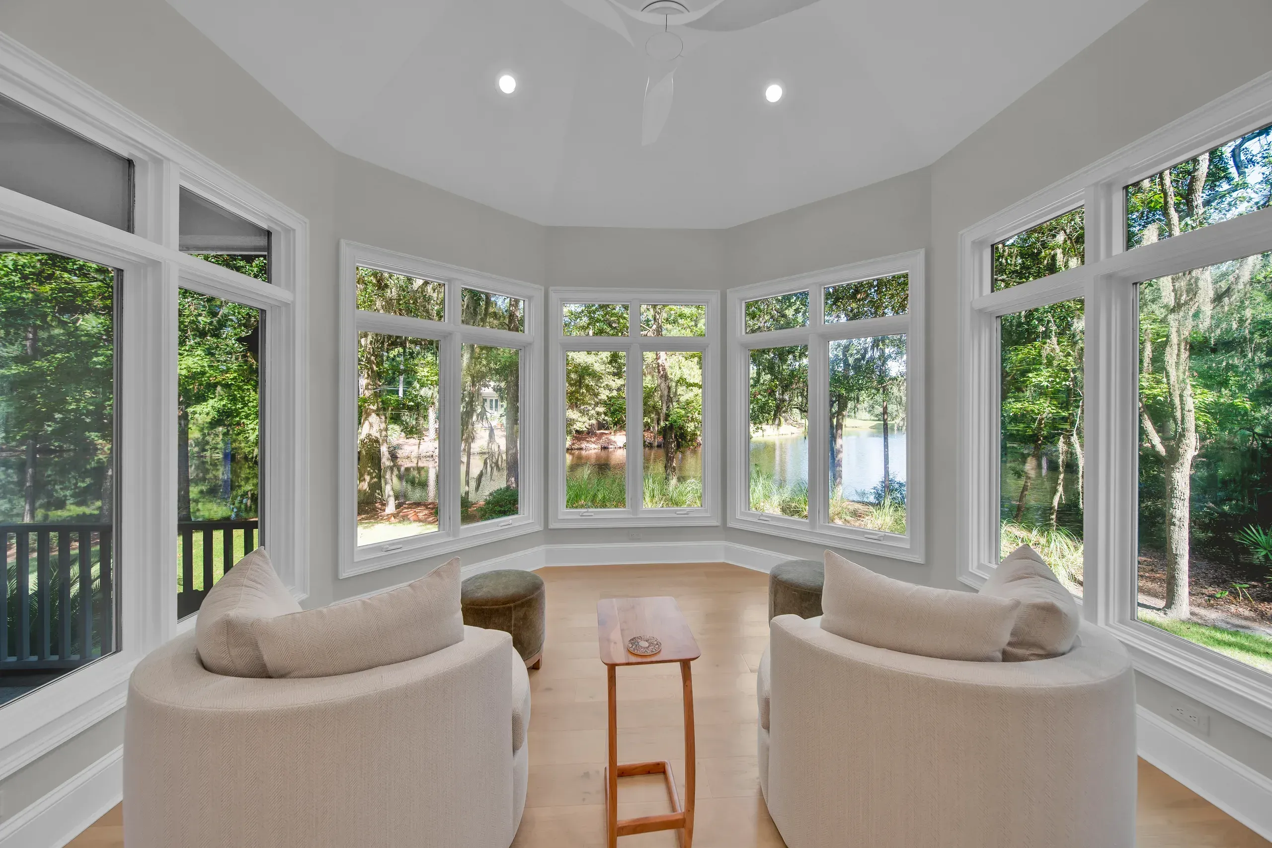 A curved sunroom with wraparound windows overlooks a wooded lagoon, furnished with two round upholstered chairs, small ottomans, and a narrow wooden side table on light hardwood floors.
