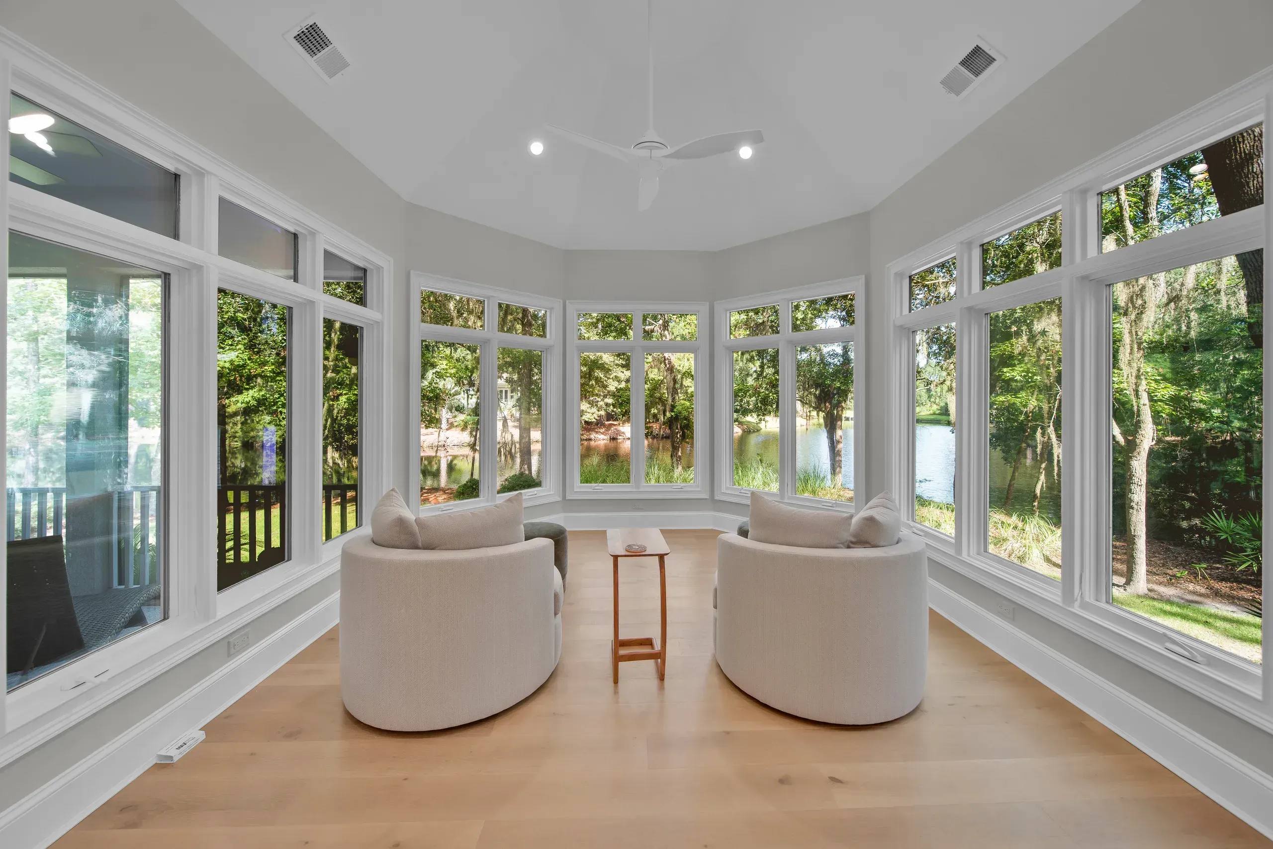 A sunroom addition with wraparound windows and light wood flooring holds two round upholstered chairs facing a small wooden side table, with a lagoon and trees visible through the glass.