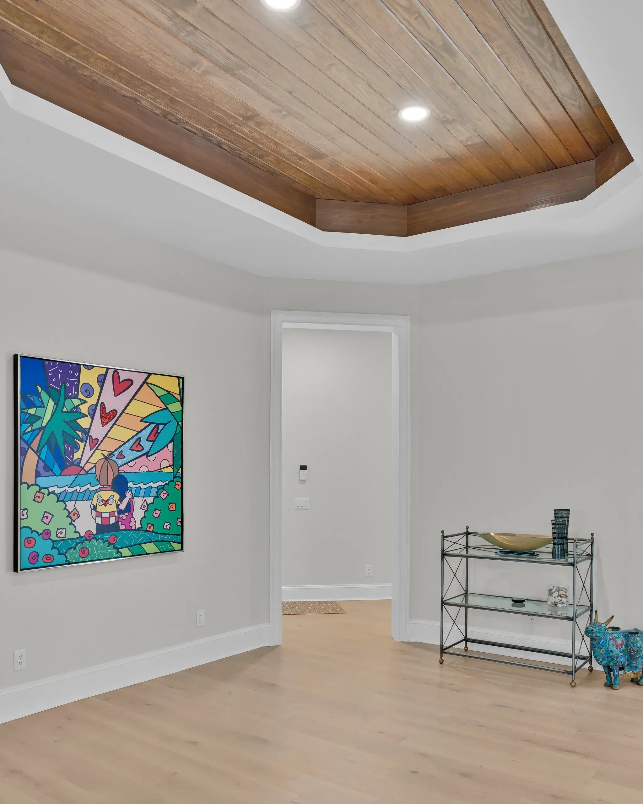 A room with a wide plank wood tray ceiling and recessed lighting features light wood flooring, a colorful abstract painting on the wall, and a metal shelving unit with decorative glass and brass accents near a doorway.