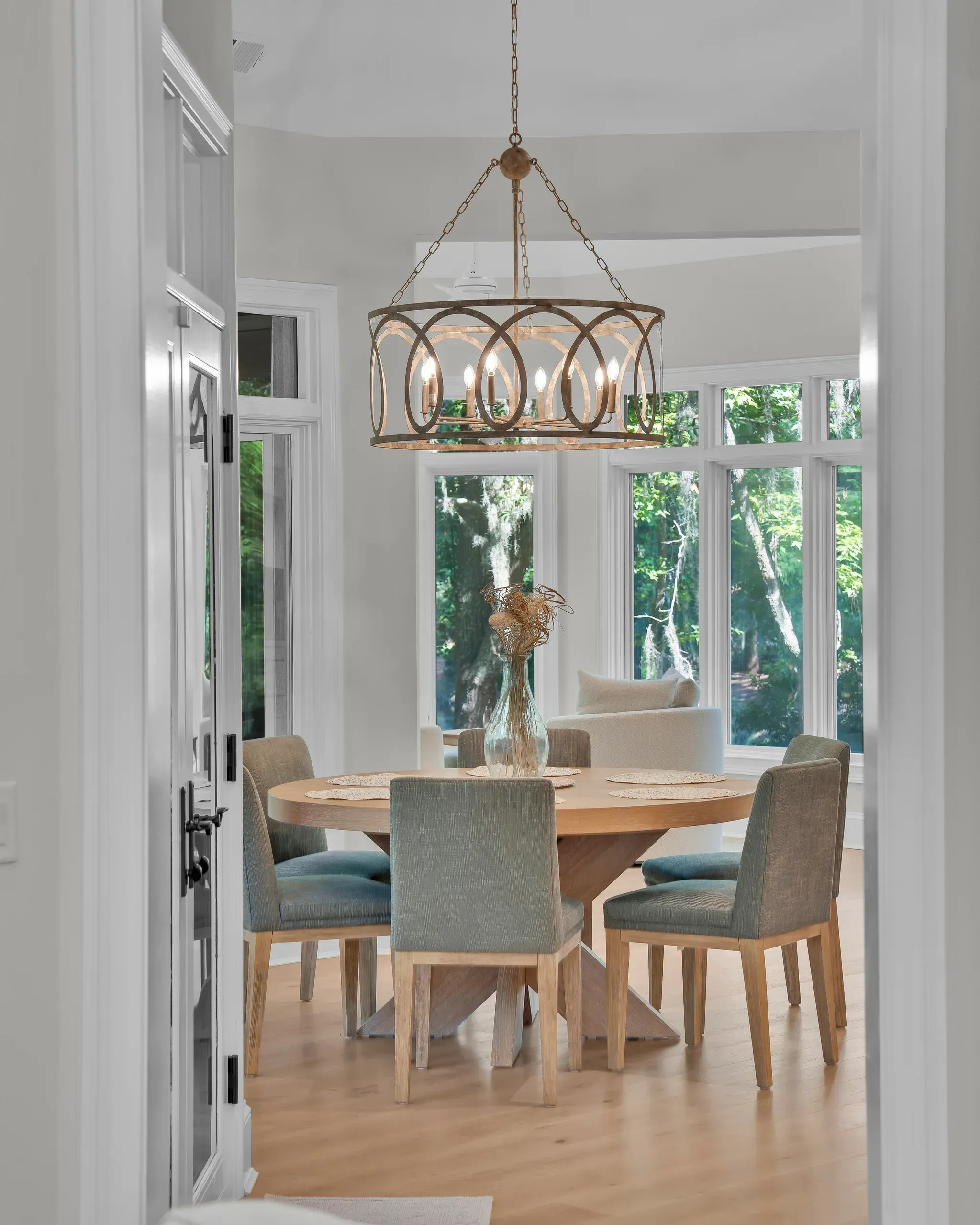 A round wood dining table surrounded by six upholstered chairs sits beneath a large drum-shaped chandelier with candle-style bulbs, framed by tall windows overlooking wooded grounds.