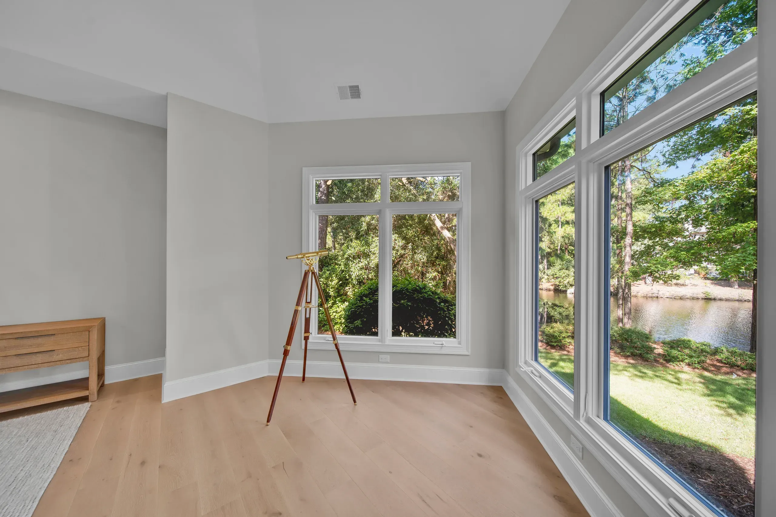 An empty room with light hardwood floors and gray walls features large corner windows overlooking a lagoon and wooded landscape, with a brass telescope on a tripod standing near a wooden console table.