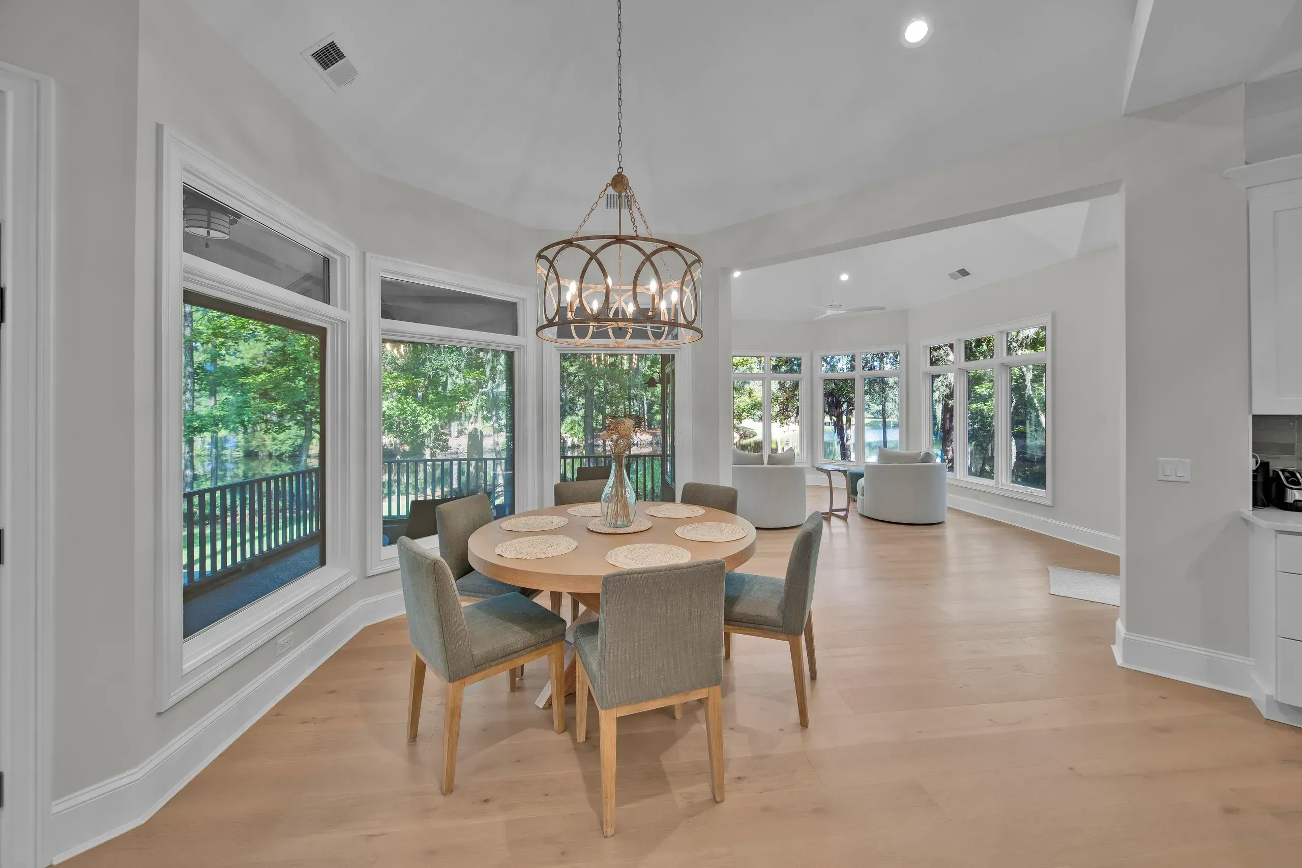 A round wooden dining table with six upholstered chairs sits beneath a circular chandelier in a sunlit bay of windows overlooking a wooded lagoon, with a connected living area and light hardwood floors extending beyond.