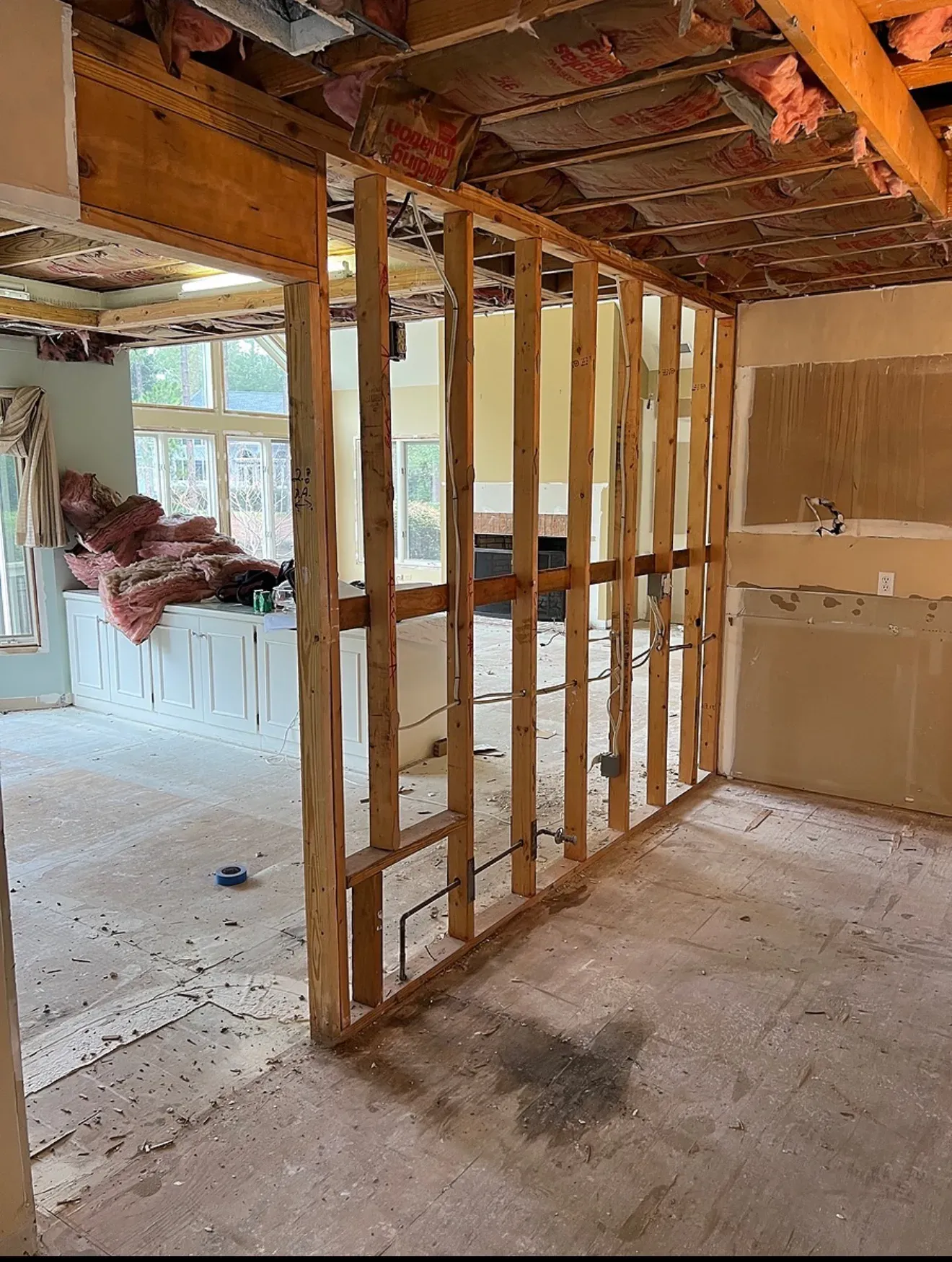 An interior under whole-home renovation shows exposed wood wall studs, ceiling joists with pink insulation, and a stripped subfloor with debris scattered throughout.