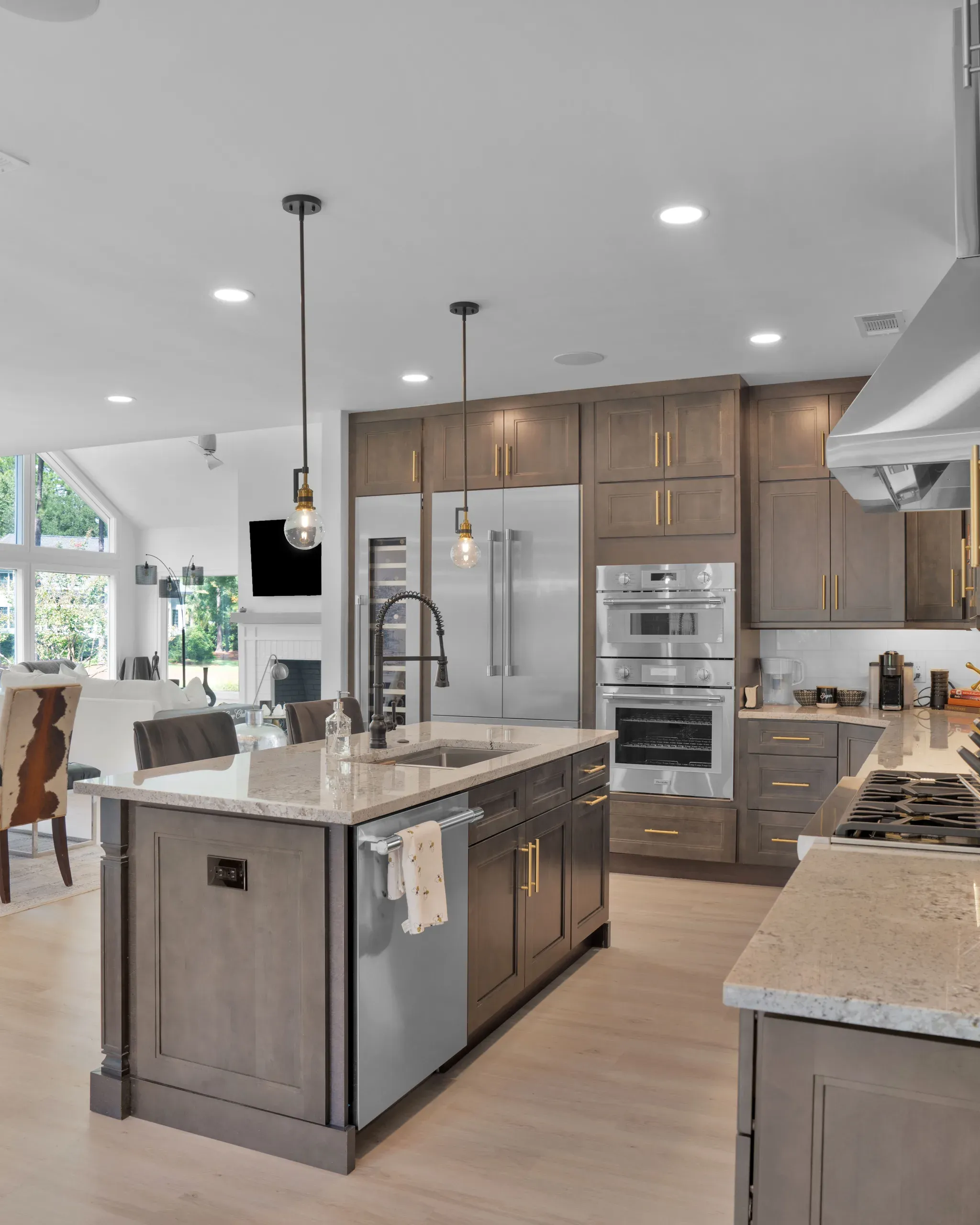 An open-concept kitchen remodel features a large island with a granite countertop, dual pendant lights, and gray cabinetry, opening onto a stainless steel refrigerator, double wall ovens, and a living area beyond.