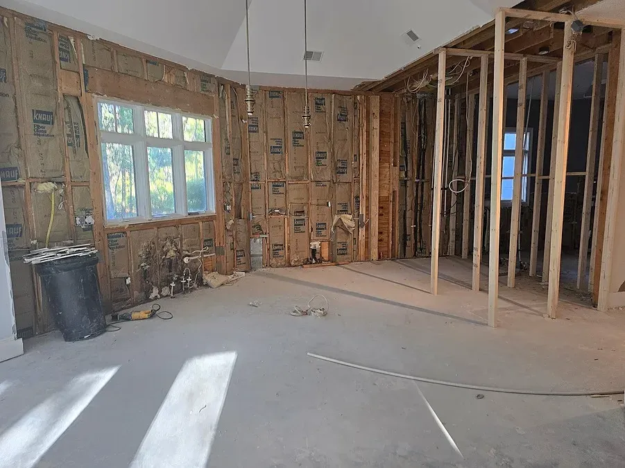 A gutted room mid-renovation shows exposed wall insulation, bare concrete flooring, dangling electrical wires, and new wood framing dividing the space near tall windows.