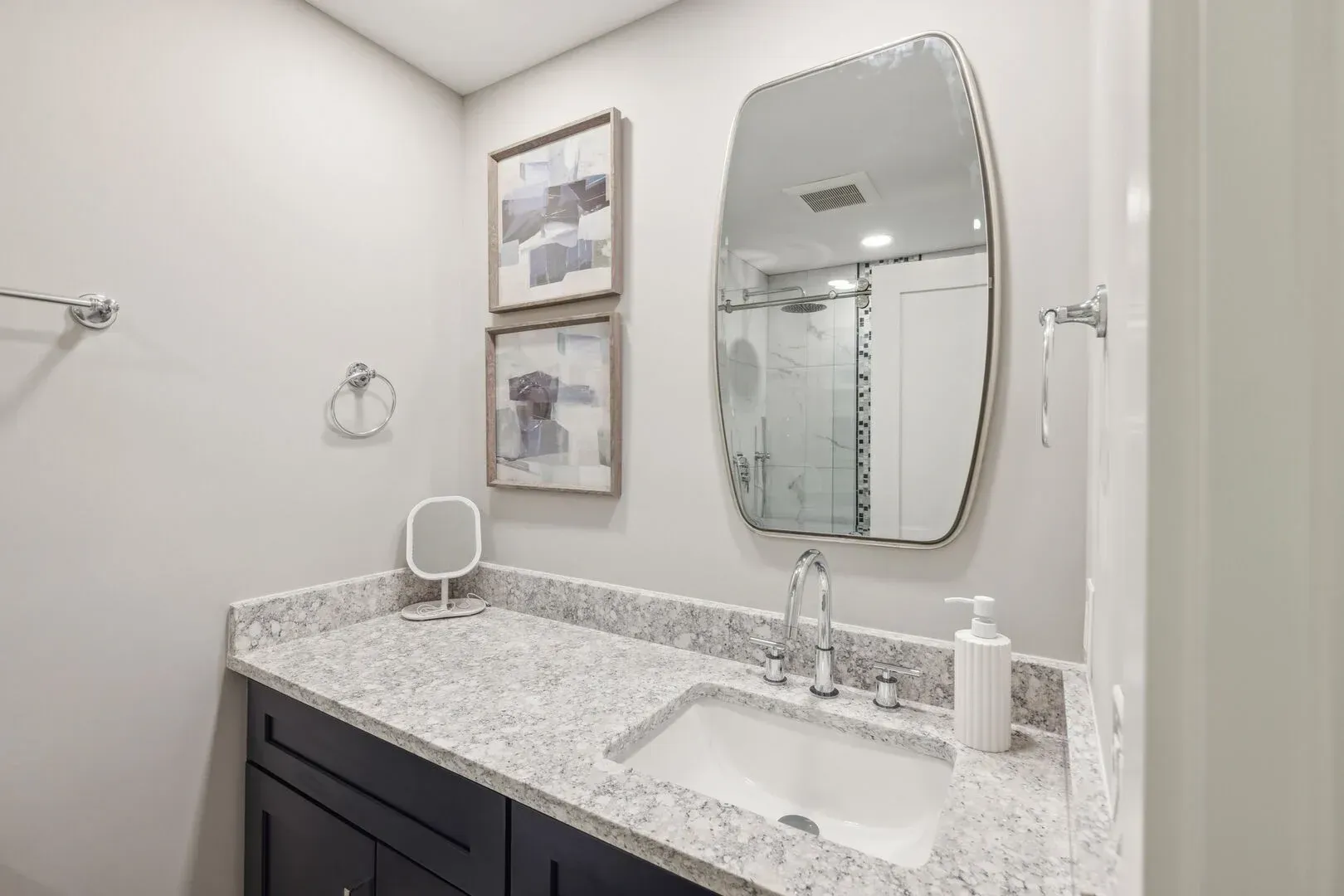 A bathroom vanity with dark cabinetry and a speckled granite countertop sits below a curved mirror that reflects a glass-enclosed shower, with framed abstract art and a small ring towel holder on the adjacent wall.