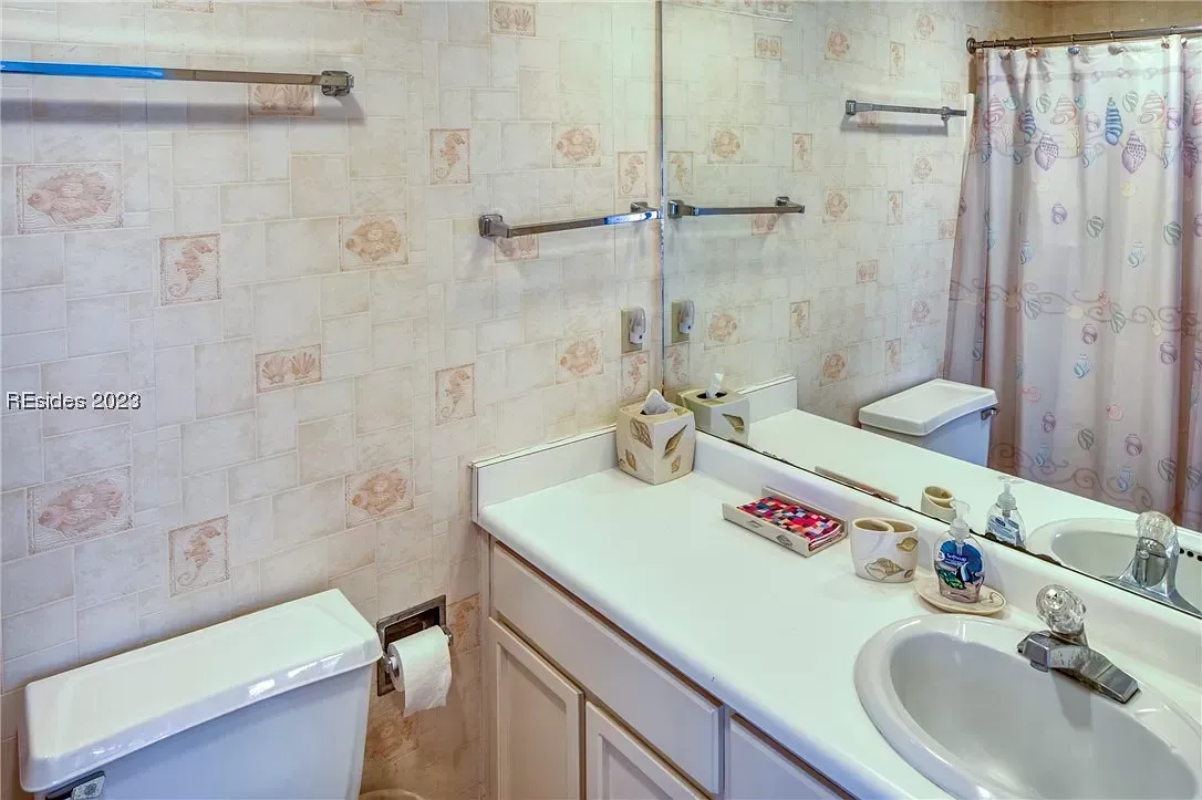 An outdated bathroom features seashell-patterned wallpaper, a white laminate vanity with double sinks, a toilet, and a shower with a coordinating fabric curtain.