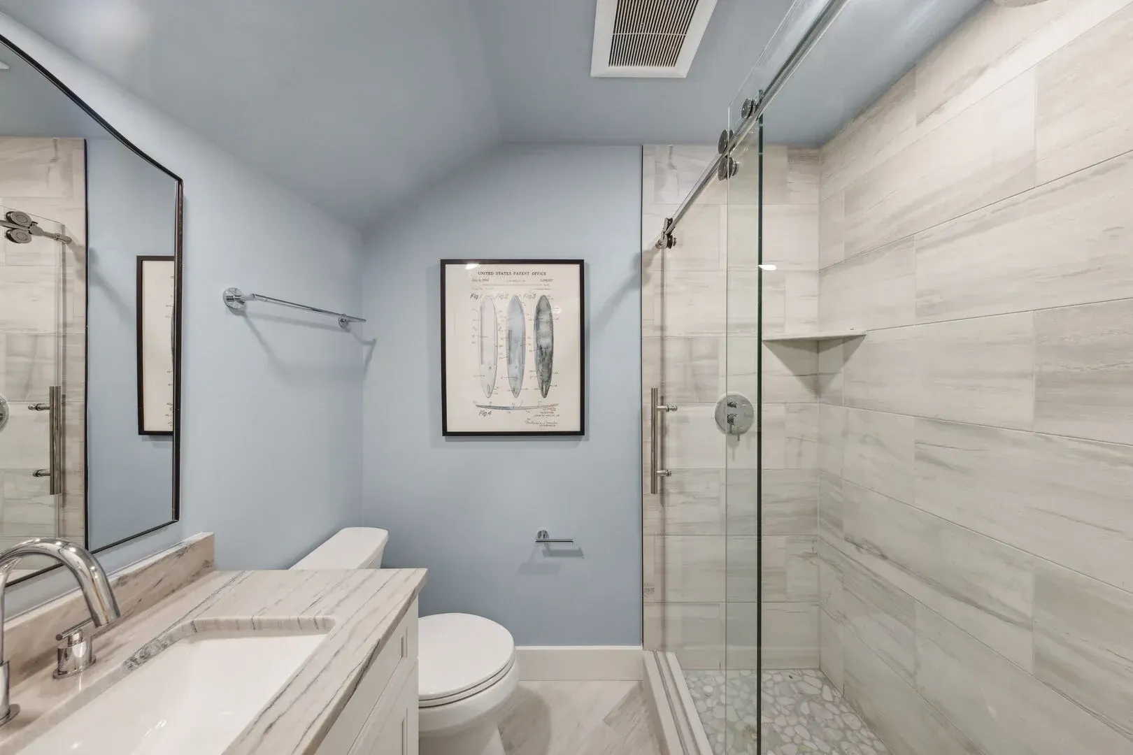 A vaulted bathroom with soft blue walls features a marble-tiled walk-in shower with a sliding glass door, a white vanity with a stone countertop, and a framed surfboard print above the toilet.