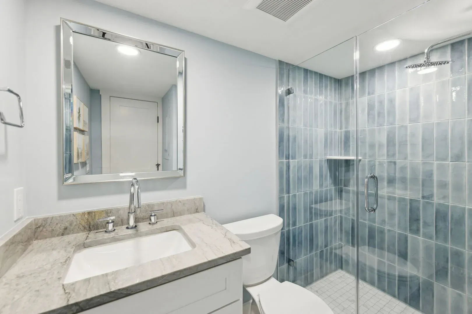 A remodeled bathroom features a marble-topped vanity with a framed mirror beside a glass-enclosed shower lined with blue vertical tile.