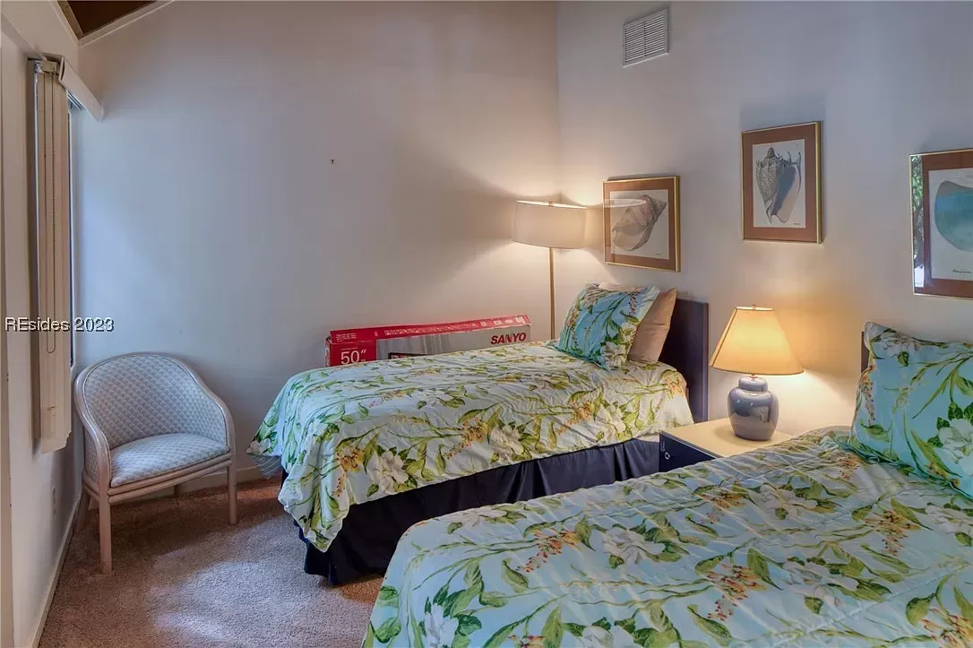 A condo guest bedroom features two twin beds with floral bedding, a bedside lamp on a nightstand, an armchair by the window, and framed shell artwork on the walls.