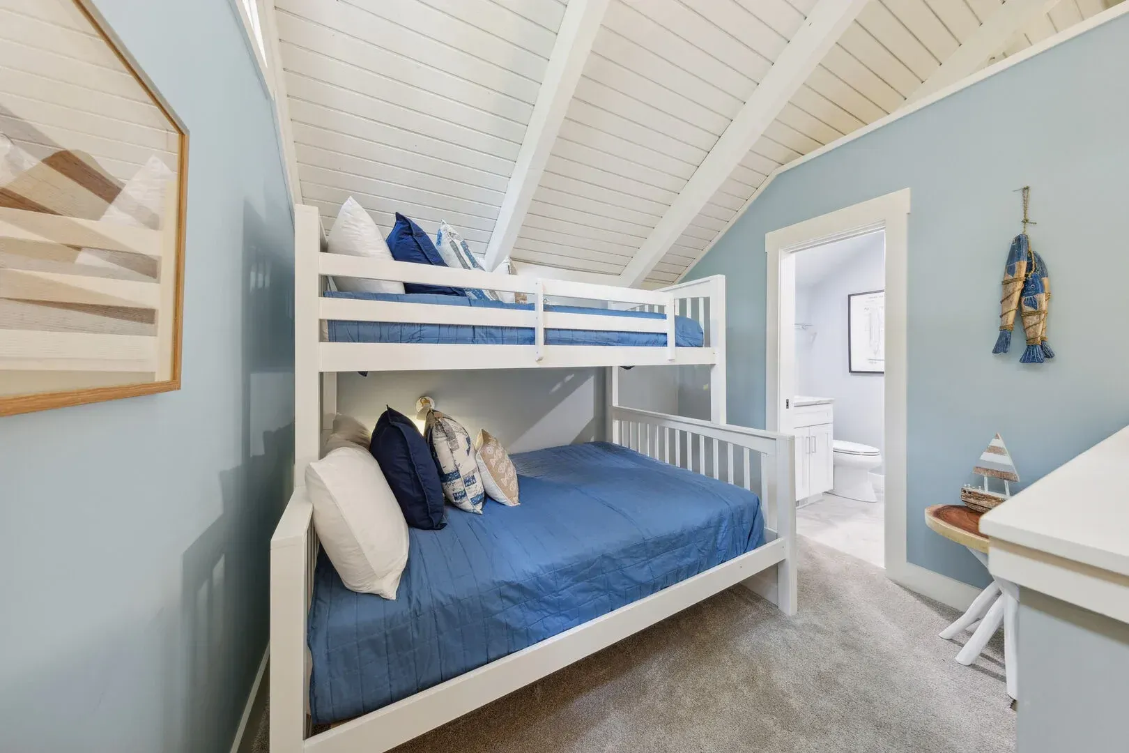 A bunk bedroom under a vaulted, shiplap-clad ceiling features a white built-in bunk bed with blue bedding, light blue walls, and a doorway leading to an attached bathroom.