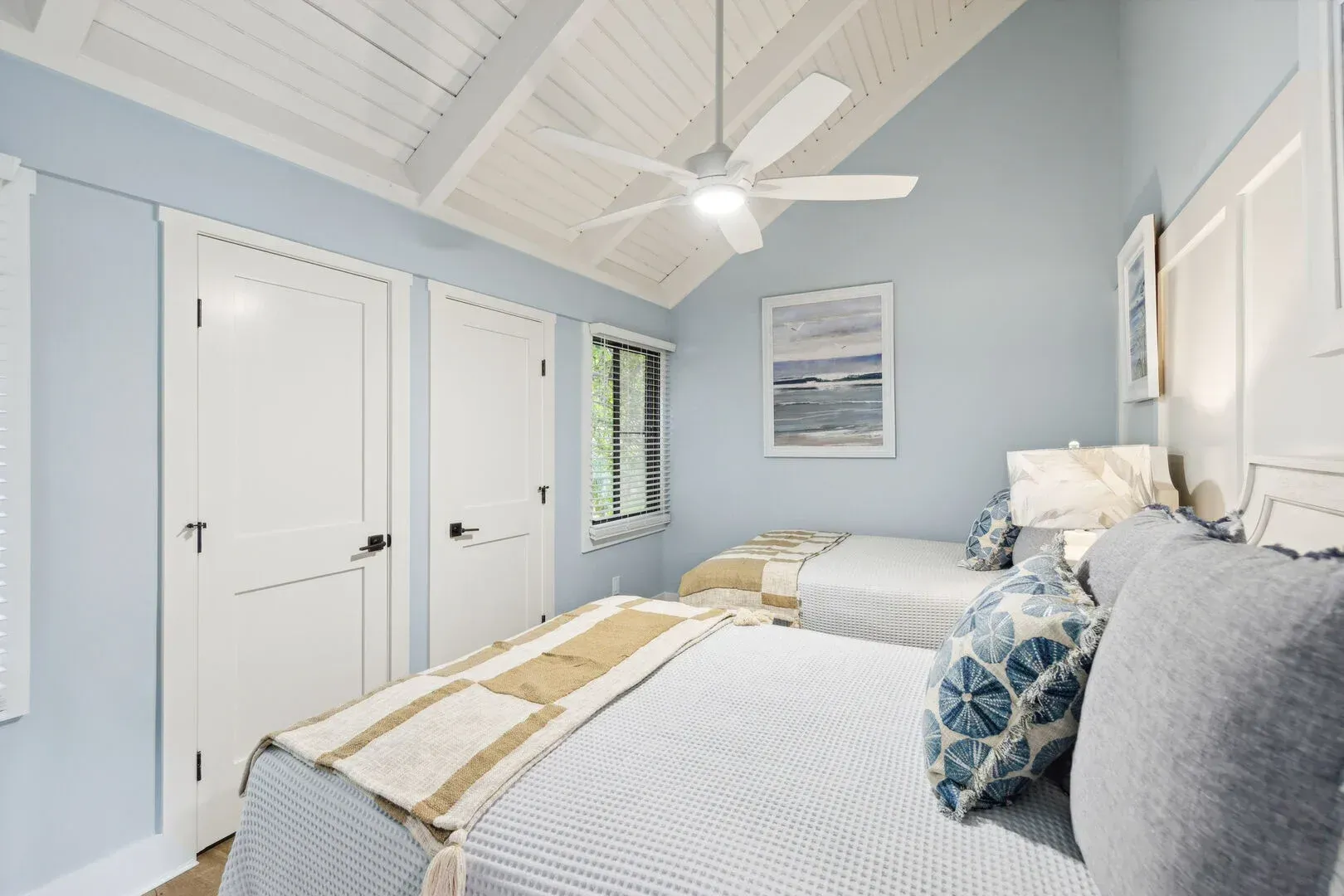 A coastal-style guest bedroom features two beds with light blue and tan bedding beneath a white vaulted shiplap ceiling with an exposed beam and ceiling fan, soft blue walls, and coastal artwork.