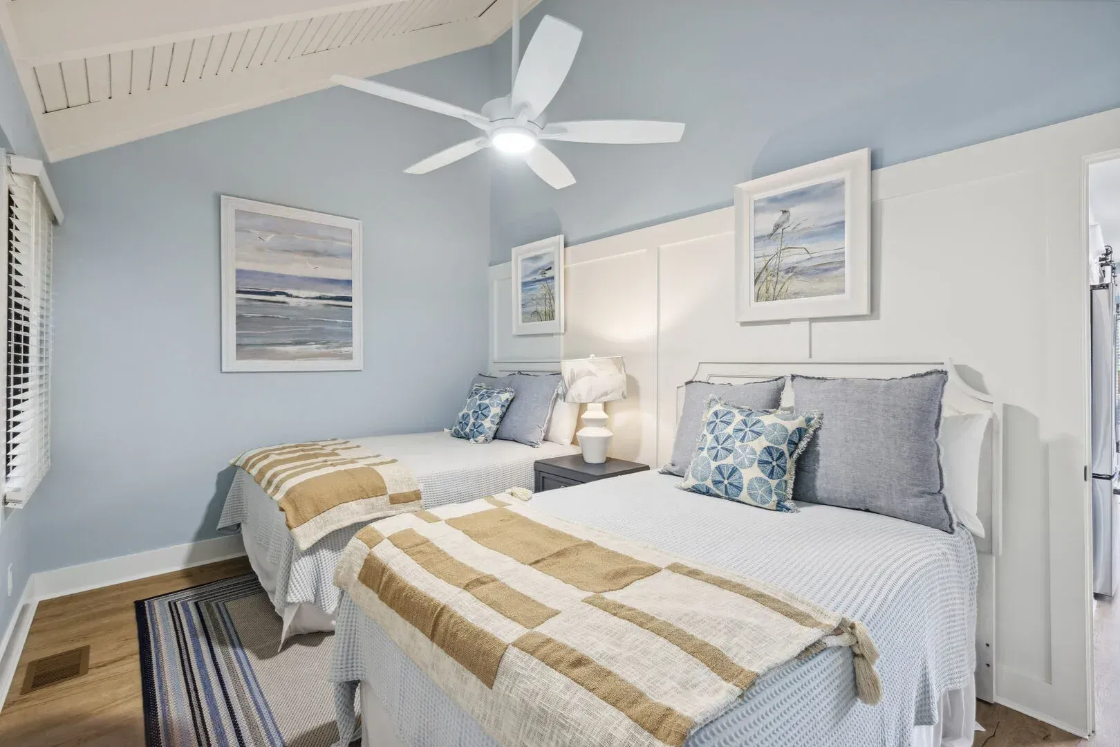 A coastal-style guest bedroom features two beds with striped tan and white throws, blue and white patterned pillows, light blue walls, white board-and-batten wainscoting, a vaulted wood-beamed ceiling with a white ceiling fan, and framed beach artwork above each headboard.
