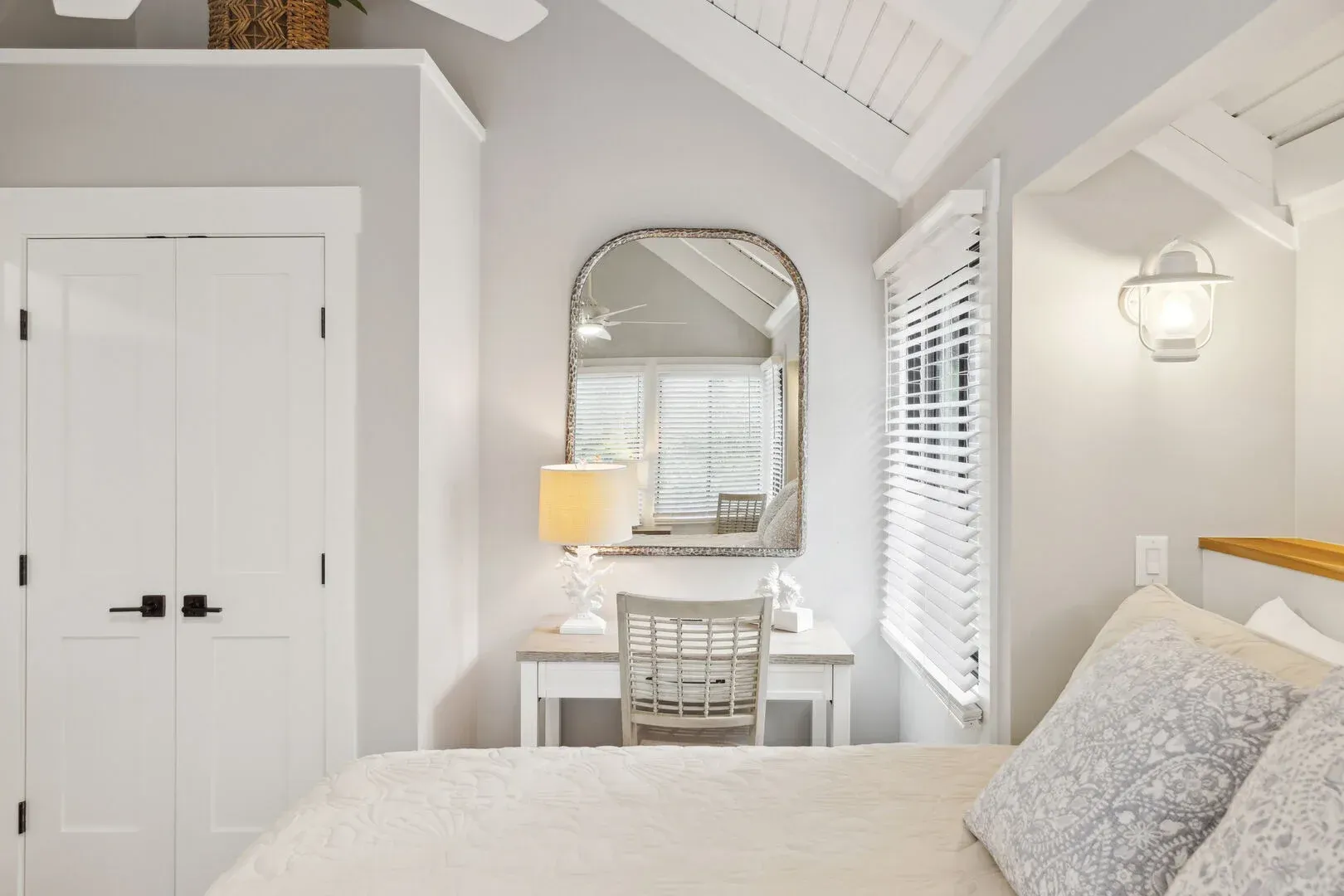 A bright bedroom with a vaulted, white-beamed ceiling features a small white desk and chair beneath an arched mirror, flanked by a wall sconce and louvered window blinds.