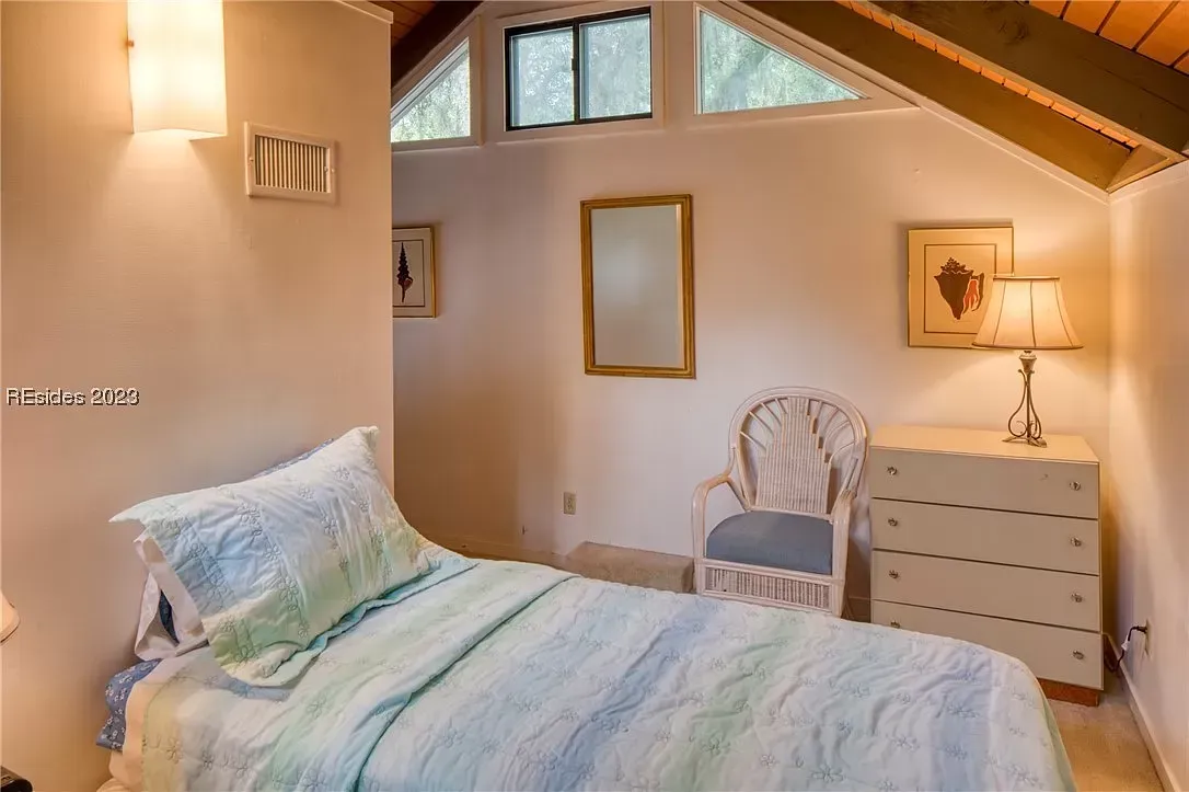 A cozy bedroom with a vaulted wood-beamed ceiling, triangular clerestory windows, a wicker accent chair, and a white dresser beside a bed with a light green quilt.