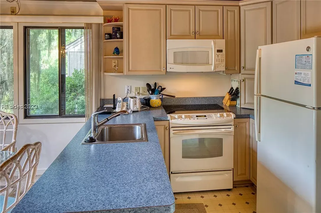 An existing kitchen with light wood cabinetry, blue speckled laminate countertops, white appliances, and a window over the sink overlooking greenery outside.