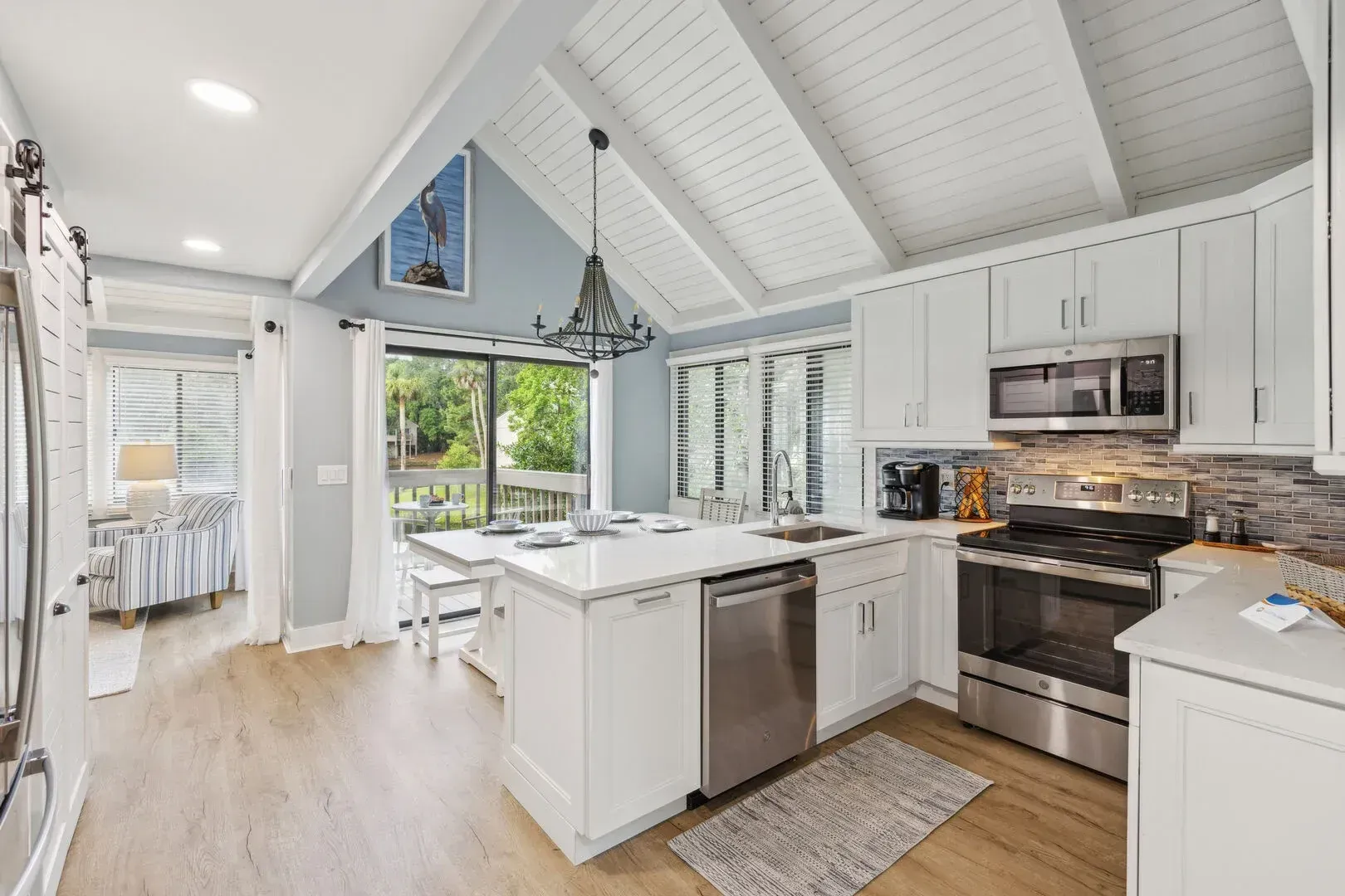 An open kitchen with white shaker cabinets, stainless steel appliances, and a white quartz island sits beneath a vaulted shiplap ceiling with exposed beams, opening into a bright sunroom with sliding glass doors leading to a deck.