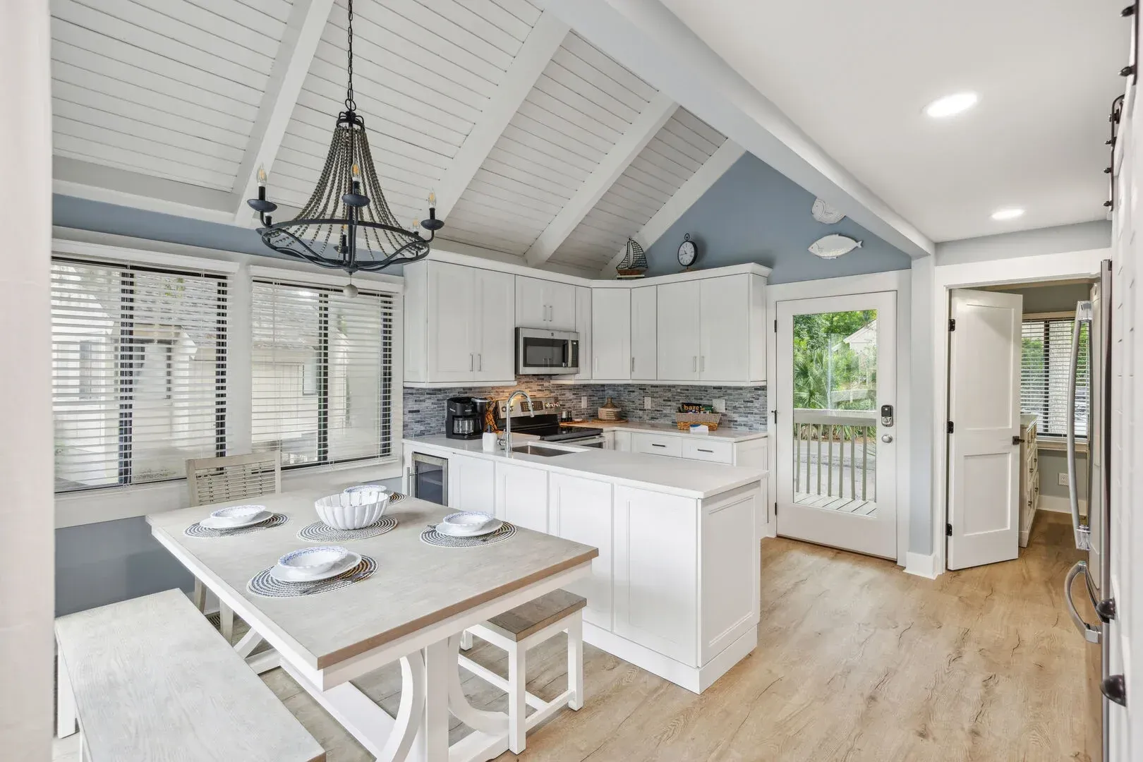 A bright coastal kitchen with white shaker cabinets, a mosaic tile backsplash, and a vaulted shiplap ceiling opens to a dining nook with a wooden table set for four.
