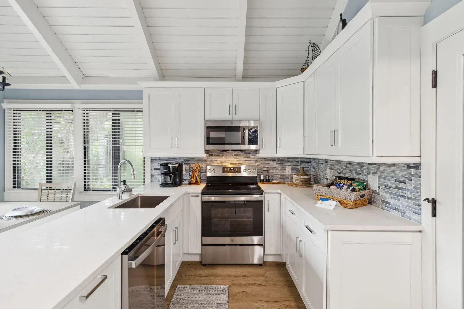 A white kitchen remodel features shaker cabinets, a stainless steel range and microwave, a mosaic tile backsplash, and a vaulted shiplap ceiling with exposed beams above light wood flooring.