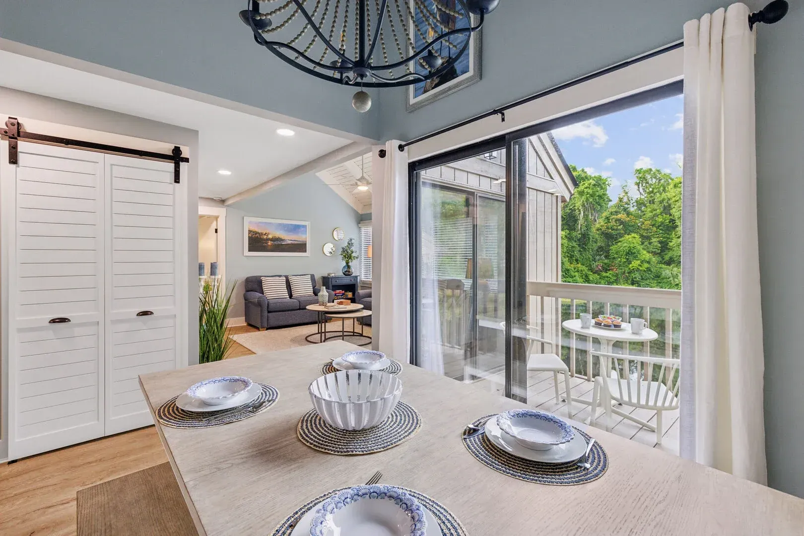A set dining table with patterned bowls sits beside a sliding glass door that opens onto a balcony overlooking green treetops, with a living room and barn-style closet doors visible in the adjoining space.