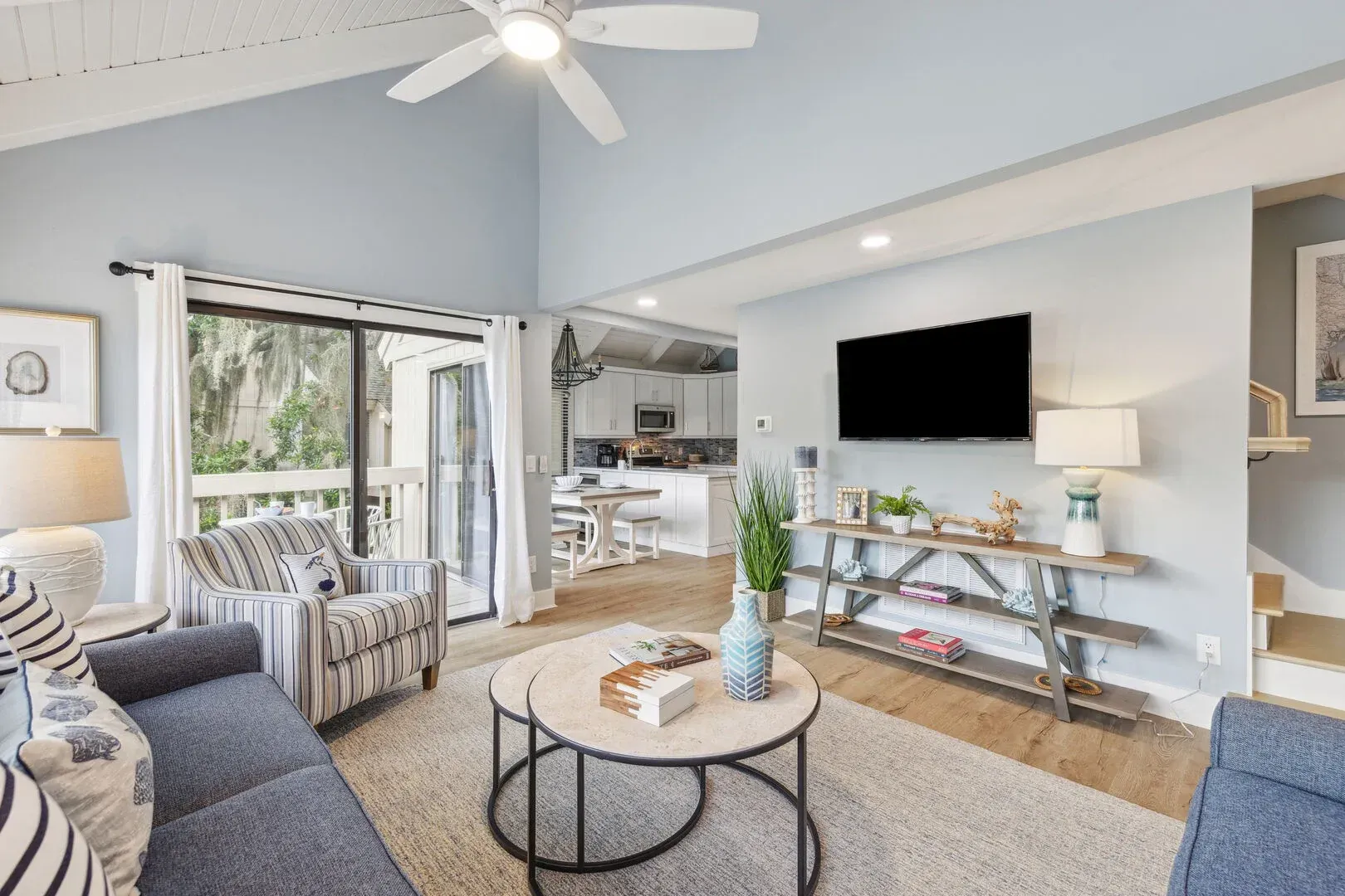 An open-concept living room with a vaulted wood ceiling and ceiling fan features blue-gray walls, a mounted flat-screen TV, and blue and striped seating around a round coffee table, with a kitchen and sliding glass door to a balcony visible beyond.