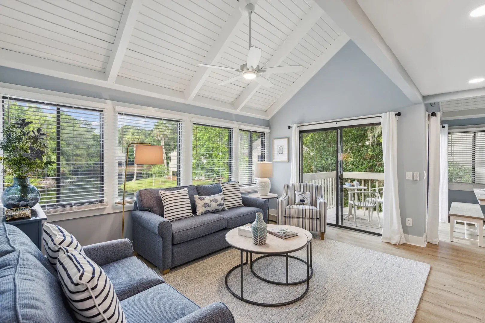 A bright living room with a vaulted, wood-planked ceiling and exposed beams features gray sofas, a round coffee table, and sliding glass doors leading to an outdoor deck.