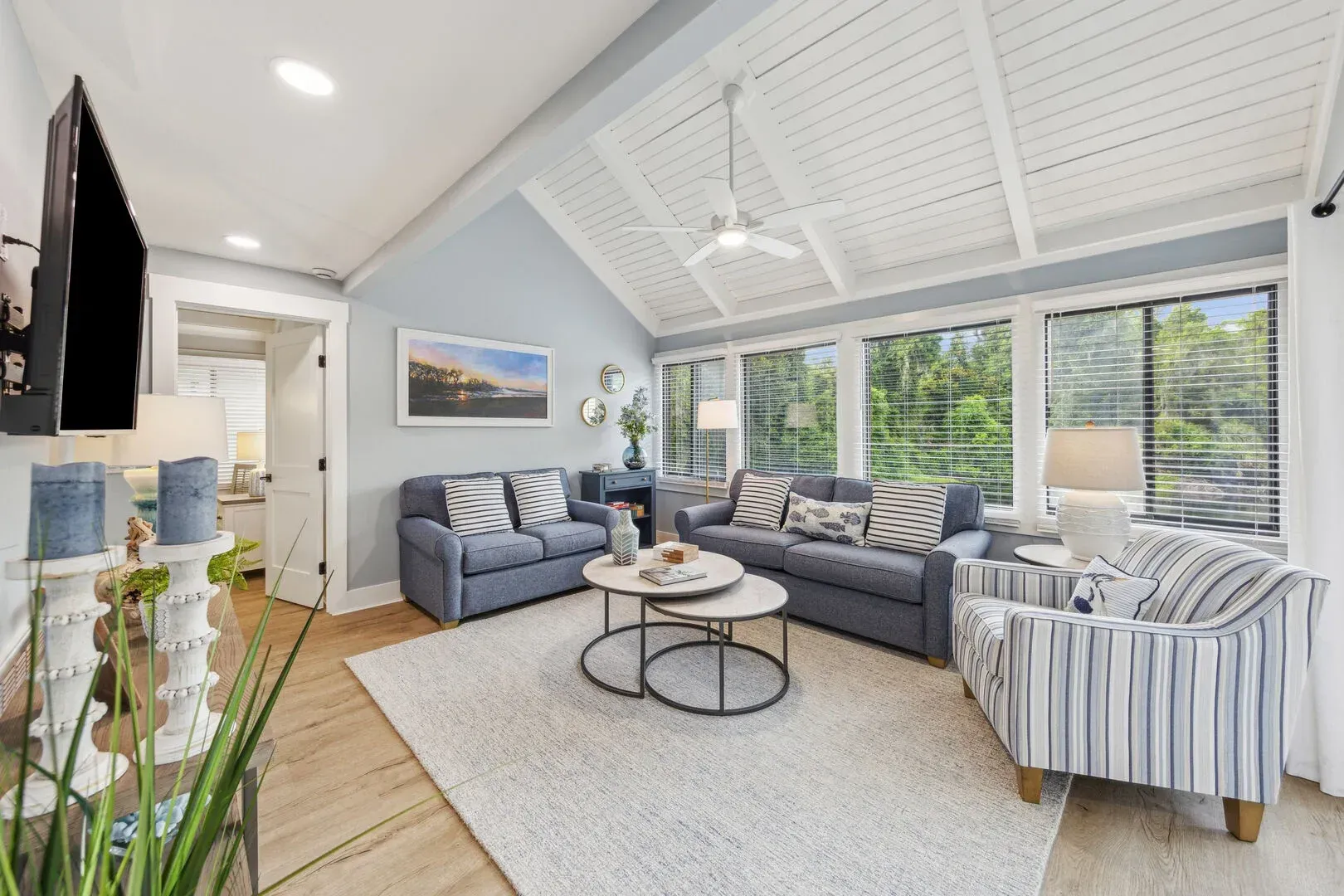 A coastal-style living room features a vaulted white beadboard ceiling with a fan, blue-gray walls, two sofas and a striped armchair around nested round coffee tables, with large windows overlooking greenery.