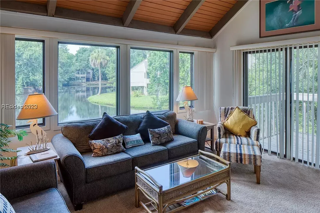 A condo living room with a vaulted wood ceiling and large windows overlooking a lagoon offers a gray sofa, striped accent chair, and glass-top coffee table on beige carpet.