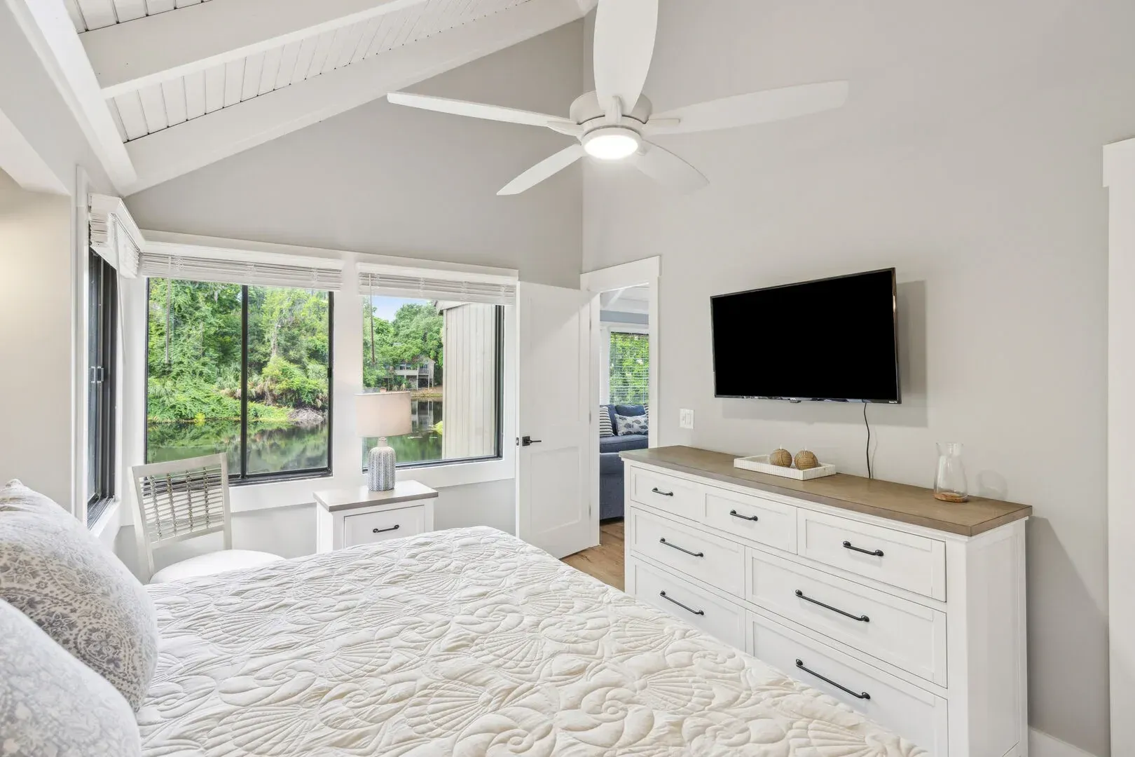 A bright primary bedroom with a vaulted, beamed ceiling features a white dresser, a wall-mounted television, and large windows overlooking a wooded lagoon view.