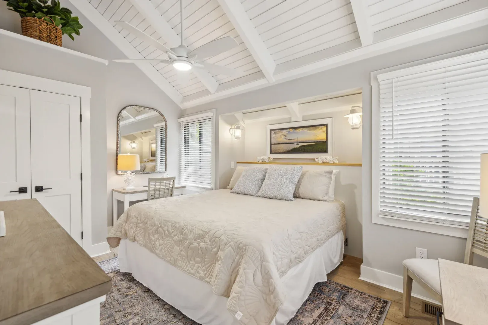 A bright primary bedroom features a vaulted wood-paneled ceiling with exposed beams, a ceiling fan, a built-in nook framing the bed with sconces, and large windows dressed in white blinds.