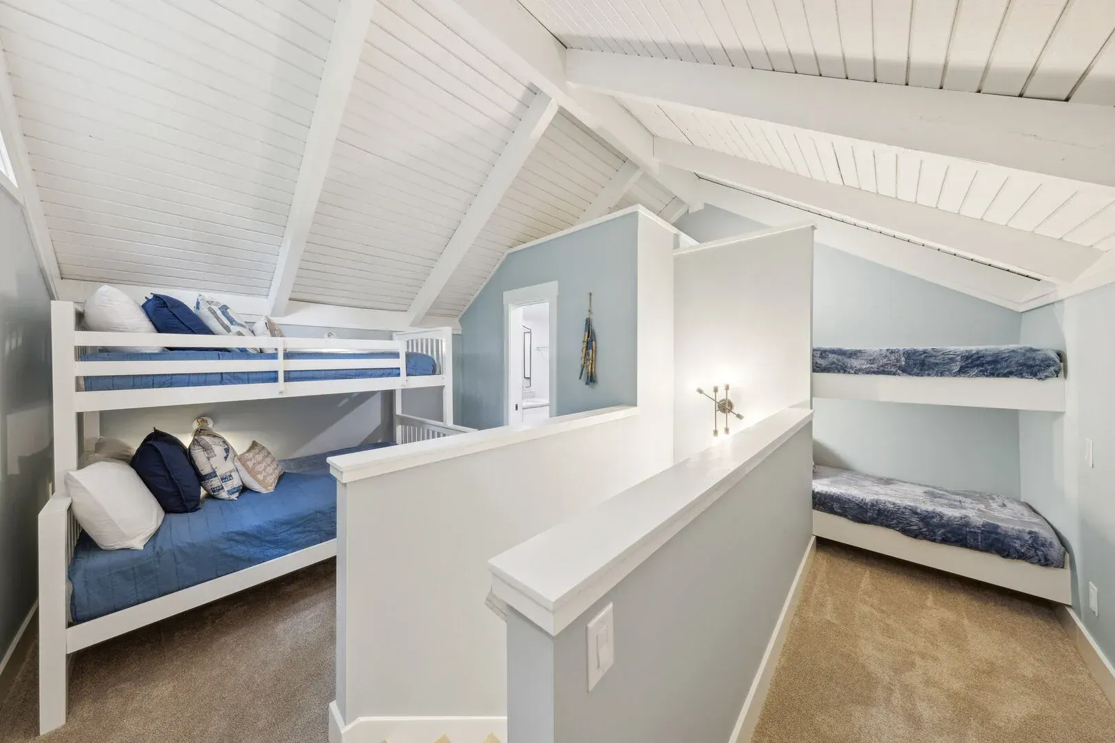 A vaulted attic bunk room with white shiplap ceilings features built-in bunk beds with blue and white bedding on both sides, separated by a low white partition wall.